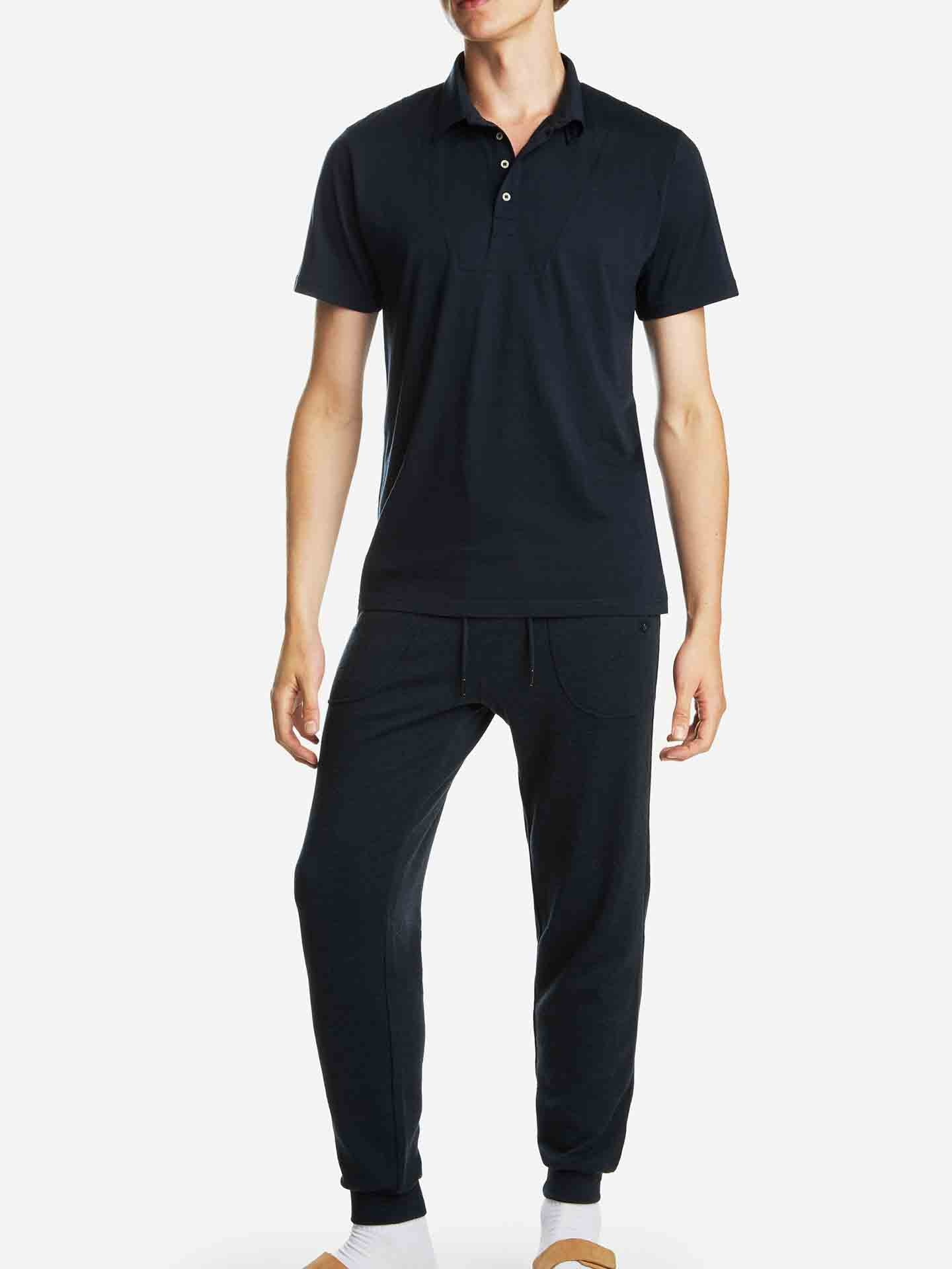 A person is standing wearing a dark navy Base One Polo Men Navy Blue shirt by We Norwegians and matching dark navy pants made from super fine merino wool. The person is also wearing white socks and tan slippers, posing against a plain white background. The outfit appears casual and comfortable.