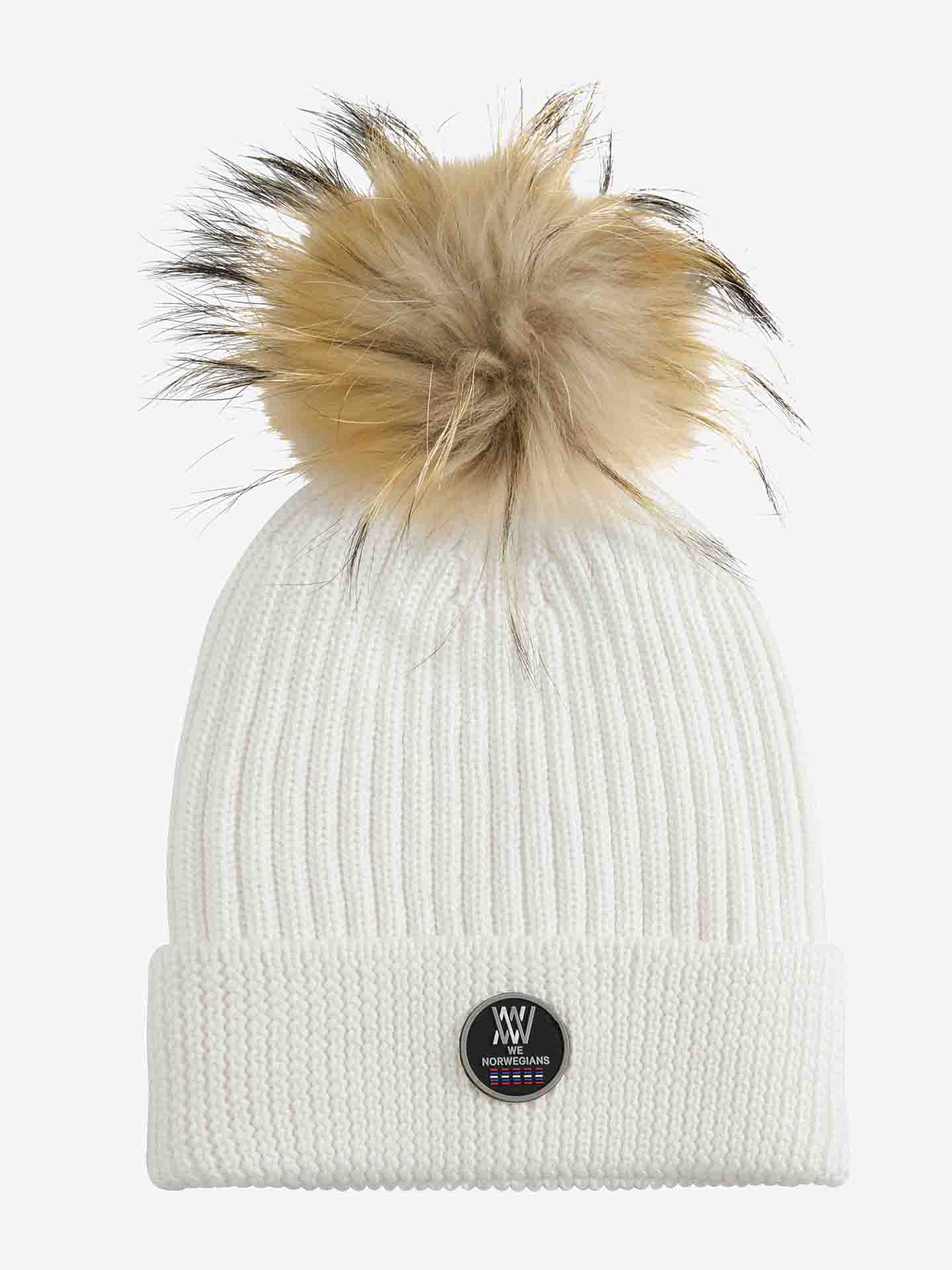 The Hemsedal Hat Women by We Norwegians is a white knitted beanie, crafted from cozy merino wool, featuring a large, fluffy tan and black pom-pom at the top. The folded brim showcases a round black patch displaying "NORWEGIANS" text, a logo, and star symbols.