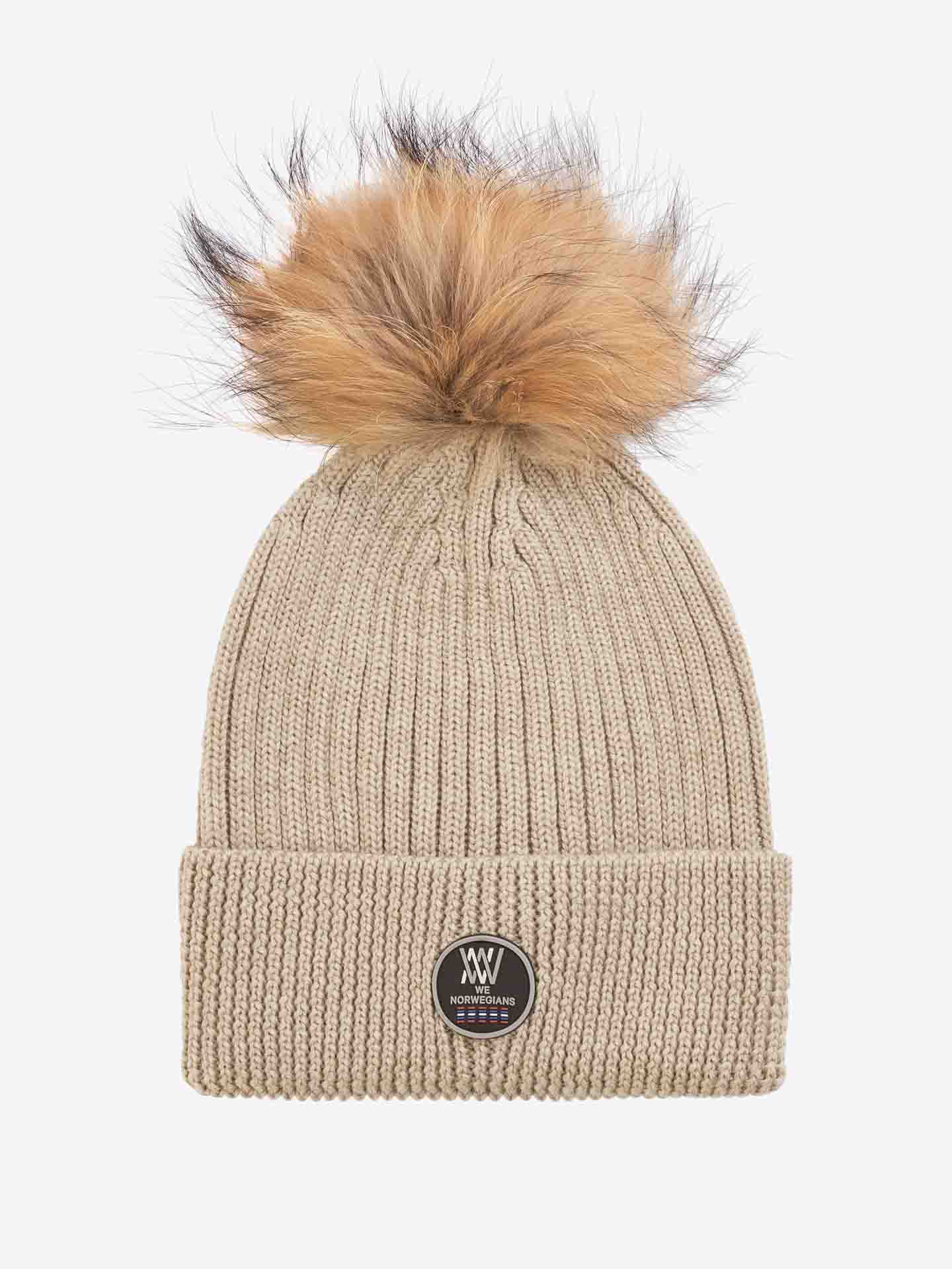 The Hemsedal Hat Women by We Norwegians is a beige, knitted beanie crafted from cozy merino wool, featuring a large, fluffy brown and black pom-pom on top. It has a folded brim and is adorned with a black circular logo patch with white text at the front.