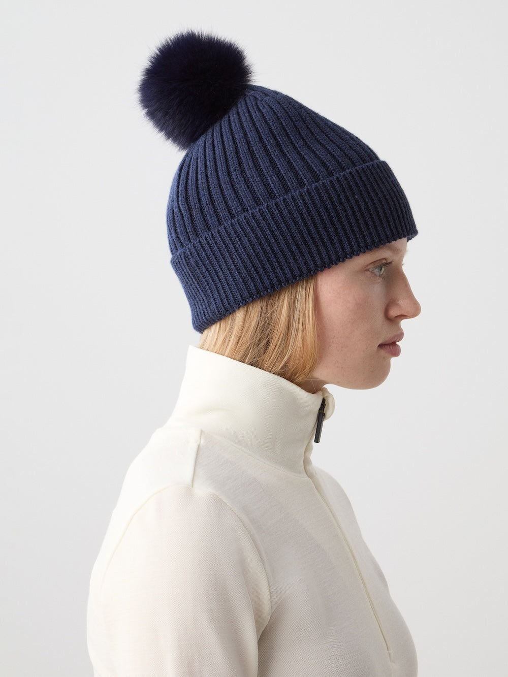 A woman in a cream zip-up top and the We Norwegians Hemsedal Hat Women—a navy blue ribbed merino wool beanie with a large dark pom-pom—stands in profile against a plain light background.