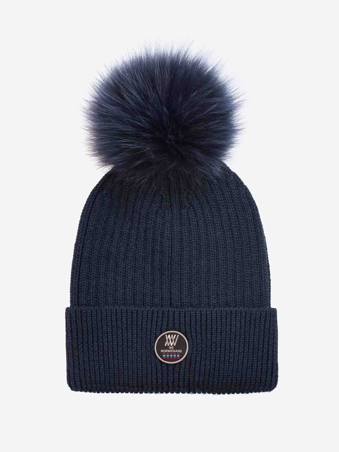 A navy blue knit Hemsedal Hat Women by We Norwegians with a folded brim and a fluffy navy pom-pom on top. The front of the beanie features a round logo patch with text and design elements in white. Made from soft merino wool, this hat is placed against a plain white background.