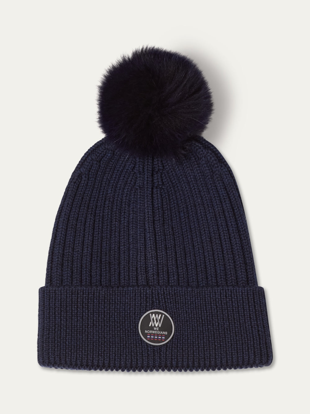 The We Norwegians Hemsedal Hat Women is a navy blue ribbed knit beanie with a large fluffy pom-pom and a folded brim, featuring a round embroidered patch, inspired by Scandinavian winter accessories.