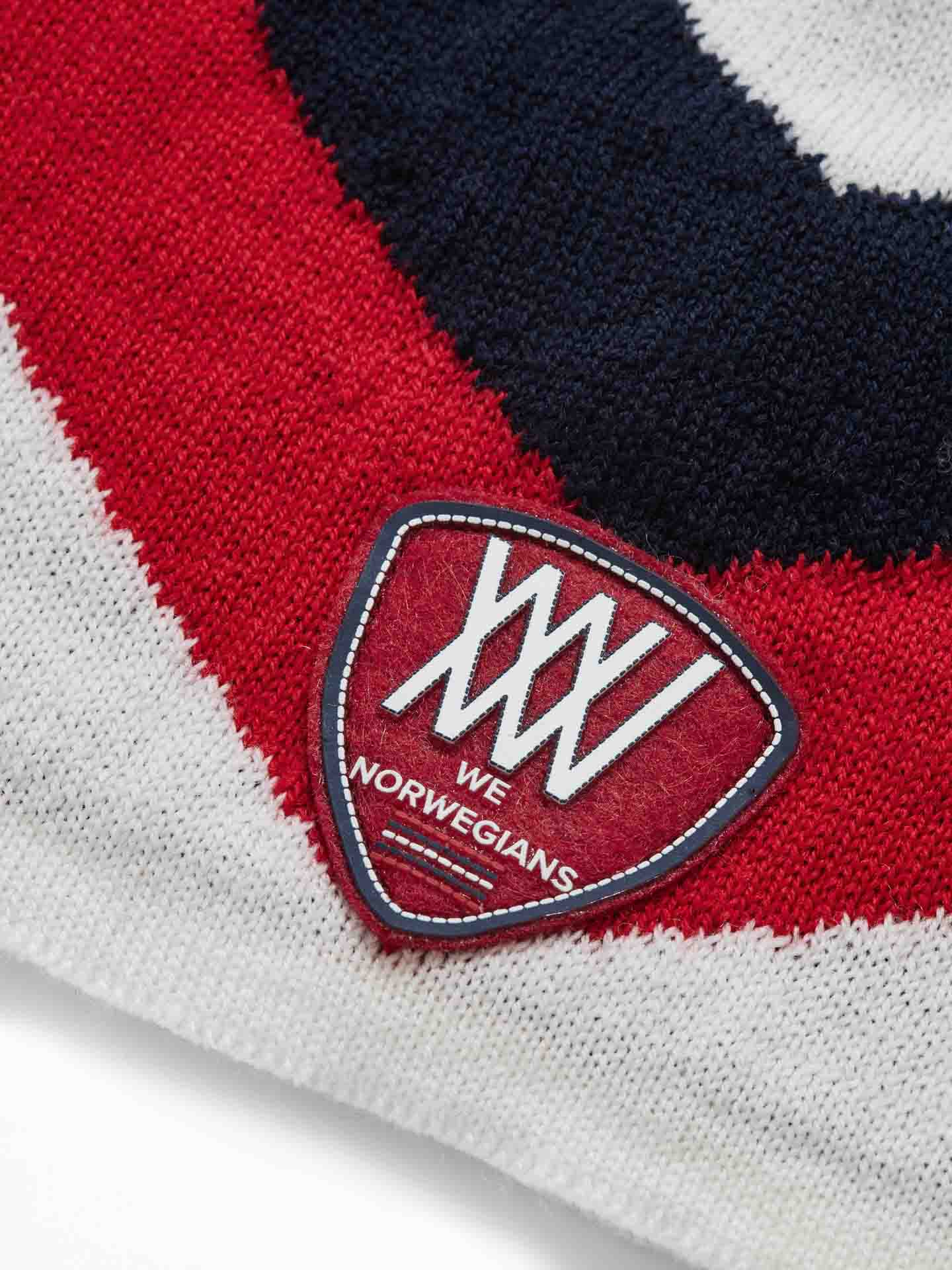 Close-up of a textile label on a garment, showcasing a red and white triangular badge with the text "We Norwegians" and stylized "W" lettering. The fabric around the label features a striking red, white, and dark blue pattern, reminiscent of the high-quality Merino wool used in the Beito Hat known for its antibacterial properties.