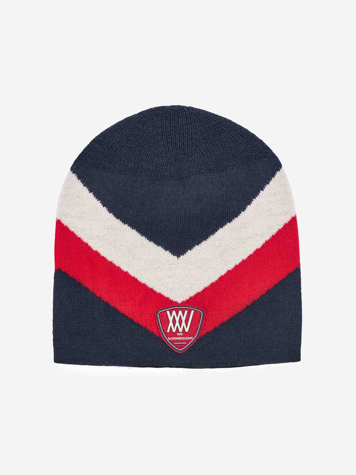 The Beito Hat by We Norwegians is a dark blue beanie featuring a white and red chevron pattern. Crafted from Merino wool, this breathable beanie includes a triangular patch at the front with the letters "WW" in white and text below it within the patch. A perfect choice for ski hat enthusiasts, the background is light gray.