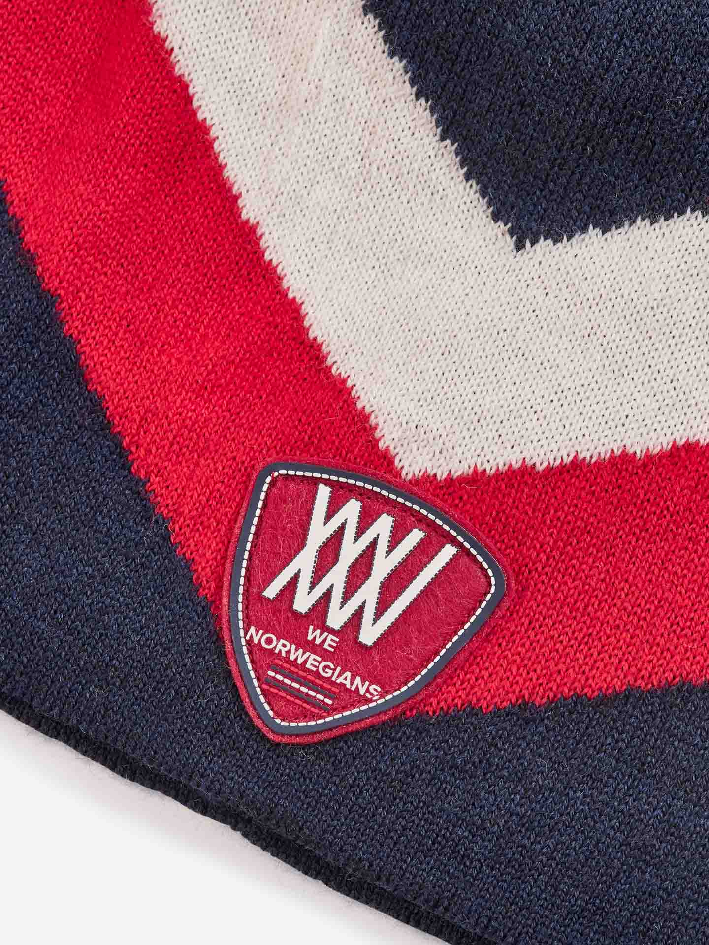 Close-up of the Beito Hat made from Merino wool, showcasing navy blue, red, and white chevron patterns. A red shield-shaped patch is sewn onto the fabric, displaying the text "XXV WE NORWEGIANS" in white lettering.