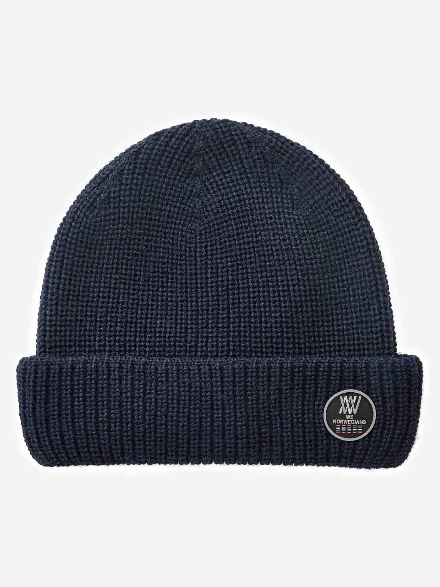 The Kvitfjell Hat from We Norwegians is a navy blue knitted beanie with a folded brim, crafted from breathable Merino wool. It features a circular patch on the front that has small text and a simple design.