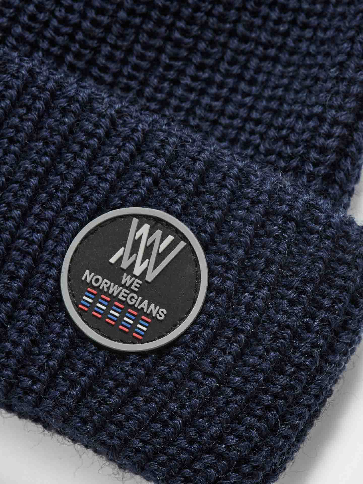 Close-up of a dark blue, waffle-knit Kvitfjell Hat crafted from breathable Merino wool. The cuff features a round, black and white patch with the "We Norwegians" logo and several small flags. The texture of the knit and the detailed patch are clearly visible.