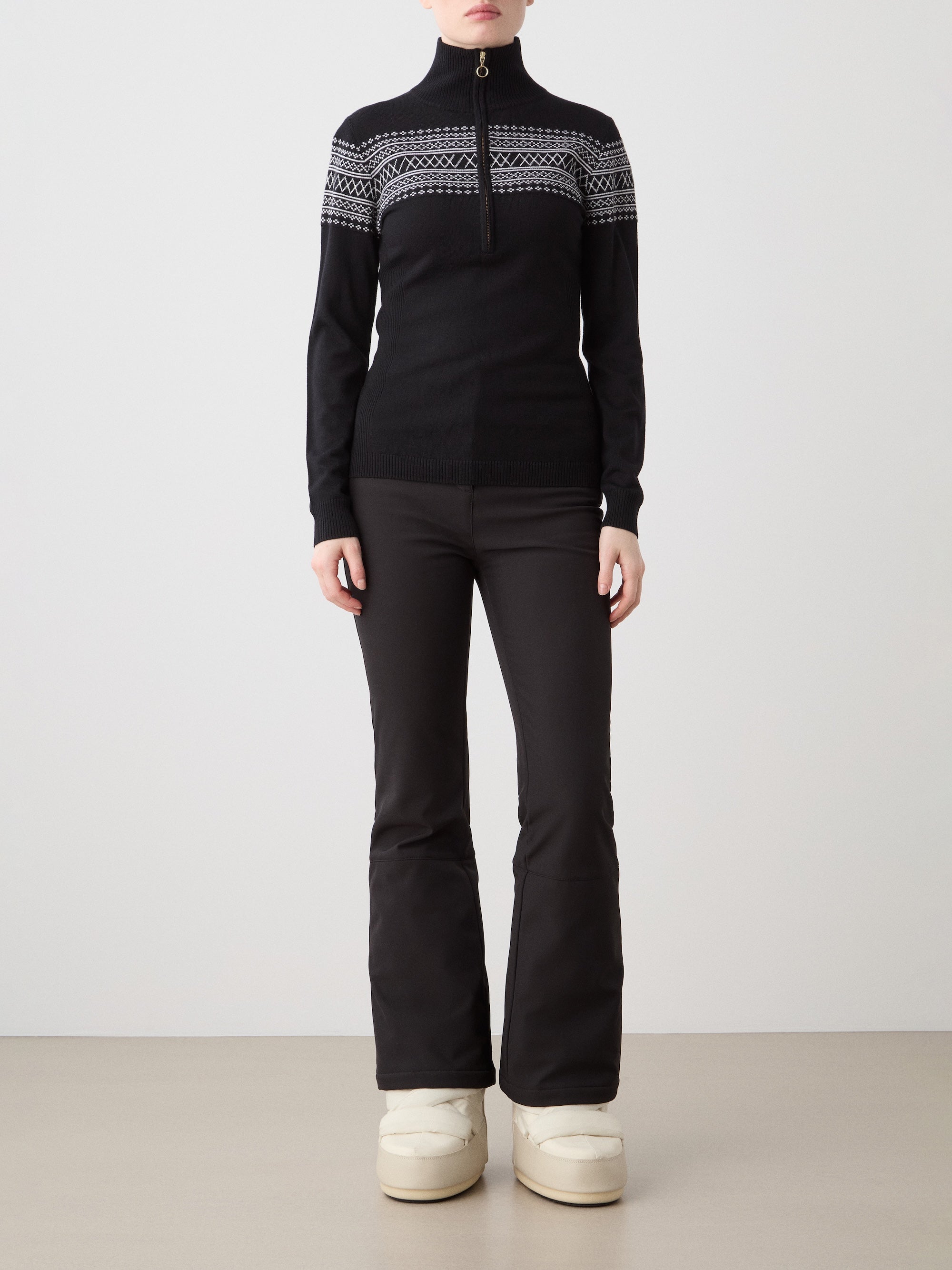 A woman stands facing forward, wearing the We Norwegians Signature Zip Up Women sweater in black and white with a Setesdal pattern, paired with black flared pants and off-white boots against a plain background.