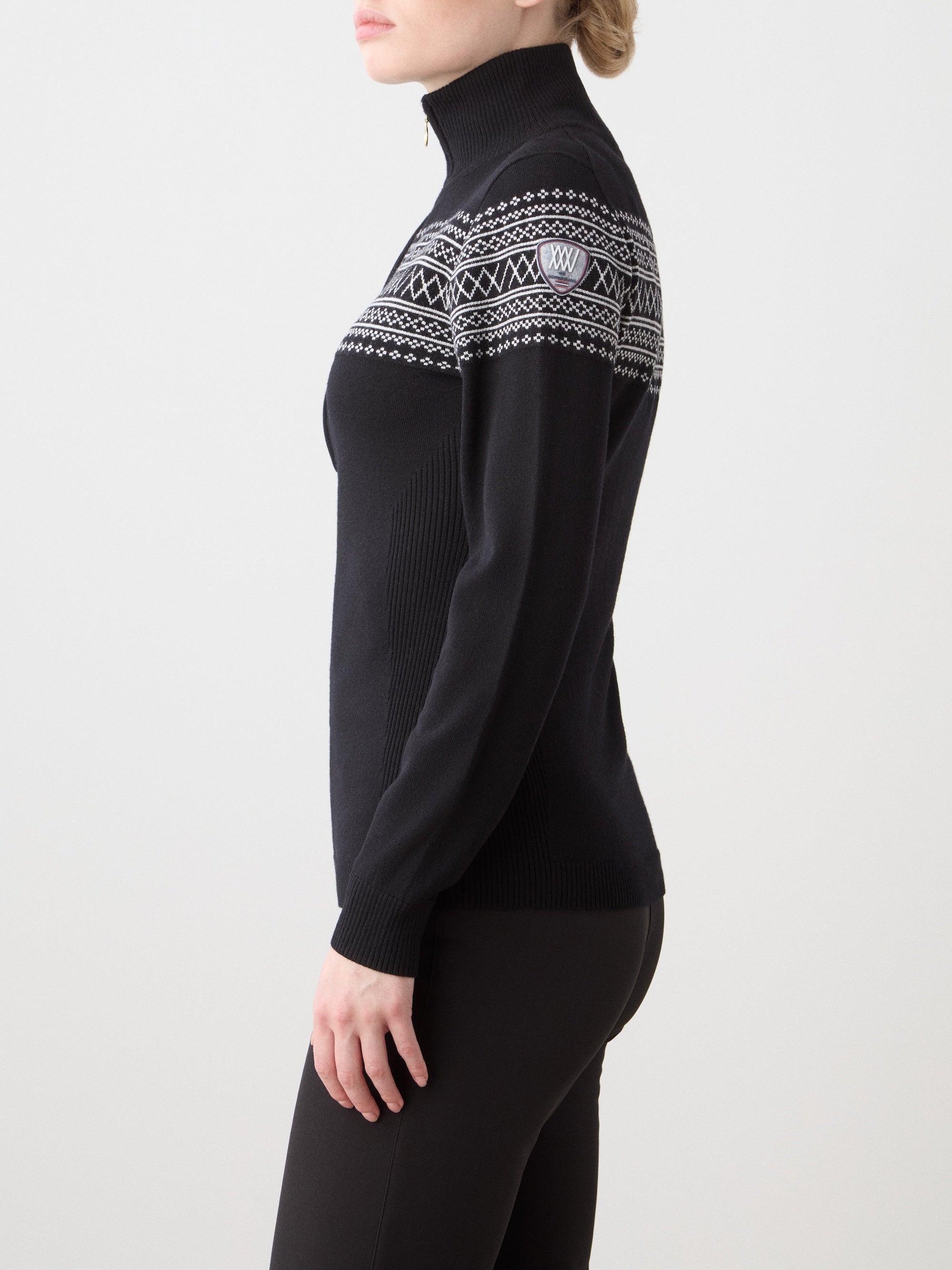 A person stands sideways in a We Norwegians Signature Zip Up Women, a black, long-sleeve, high-neck sweater with a white Setesdal pattern on the chest and shoulders, made from washable merino yarn, paired with black pants. The background is light and plain.