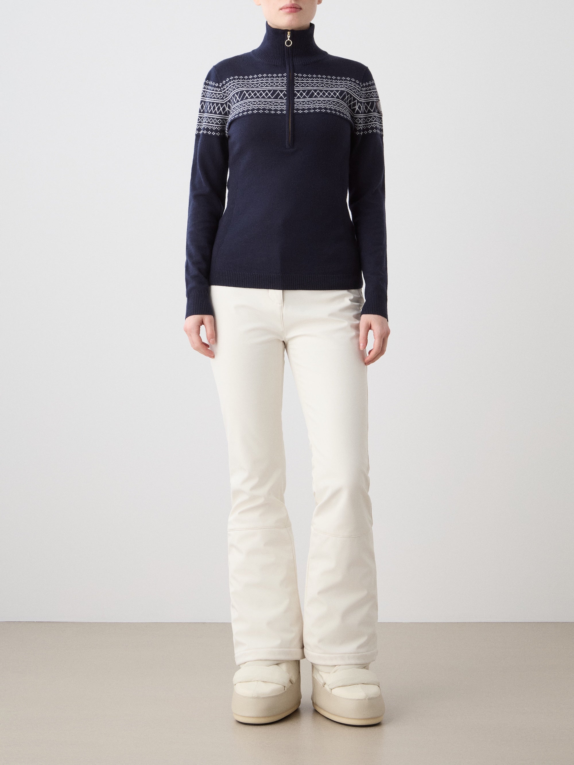 A person stands facing forward wearing We Norwegians' Signature Zip Up Women in navy blue with a classic Setesdal pattern, paired with off-white pants and beige snow boots against a plain background.