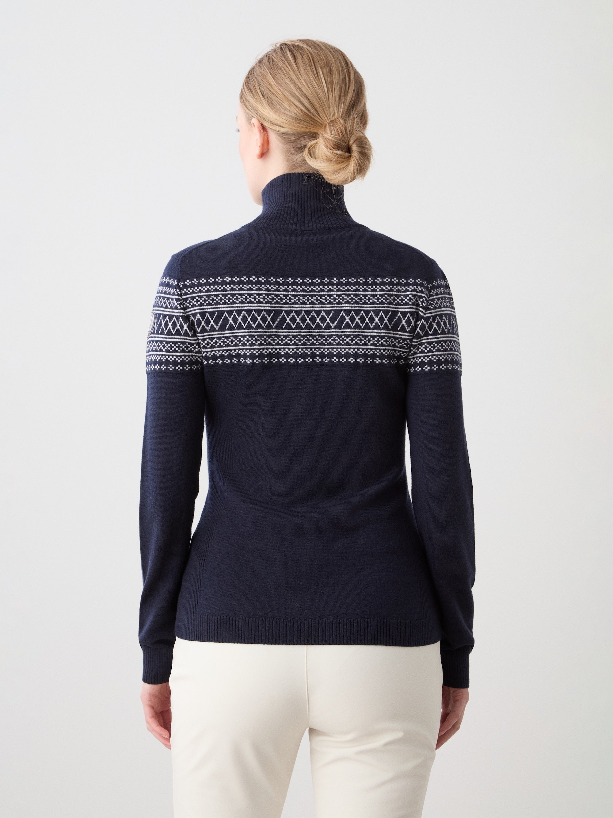A woman with blonde hair in a low bun models the We Norwegians Signature Zip Up Women—a navy merino wool sweater with a white Setesdal pattern on the shoulders and sleeves—paired with light-colored pants, shown from the back against a plain background.