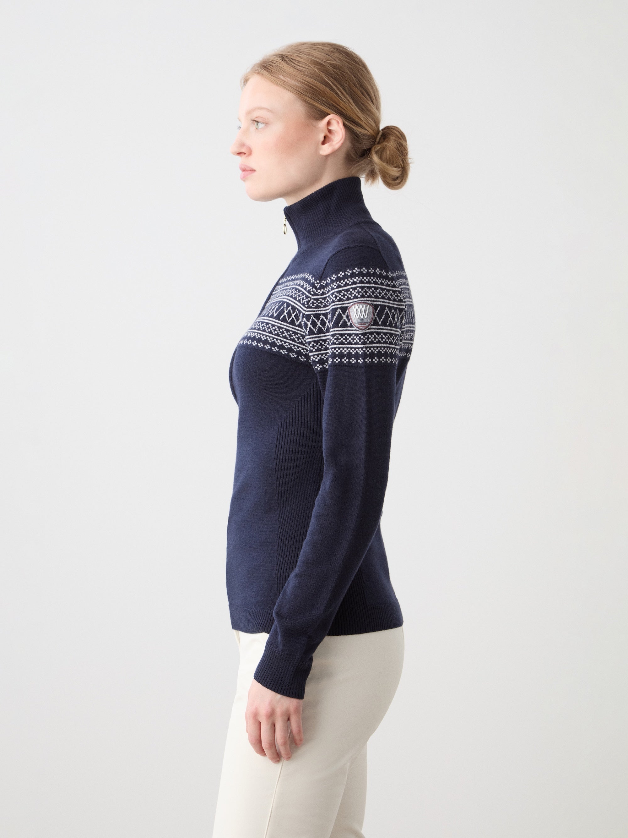 A woman with light skin and blonde hair in a low bun wears the We Norwegians Signature Zip Up Women sweater in navy with Setesdal pattern and white pants, standing in profile against a plain light background.