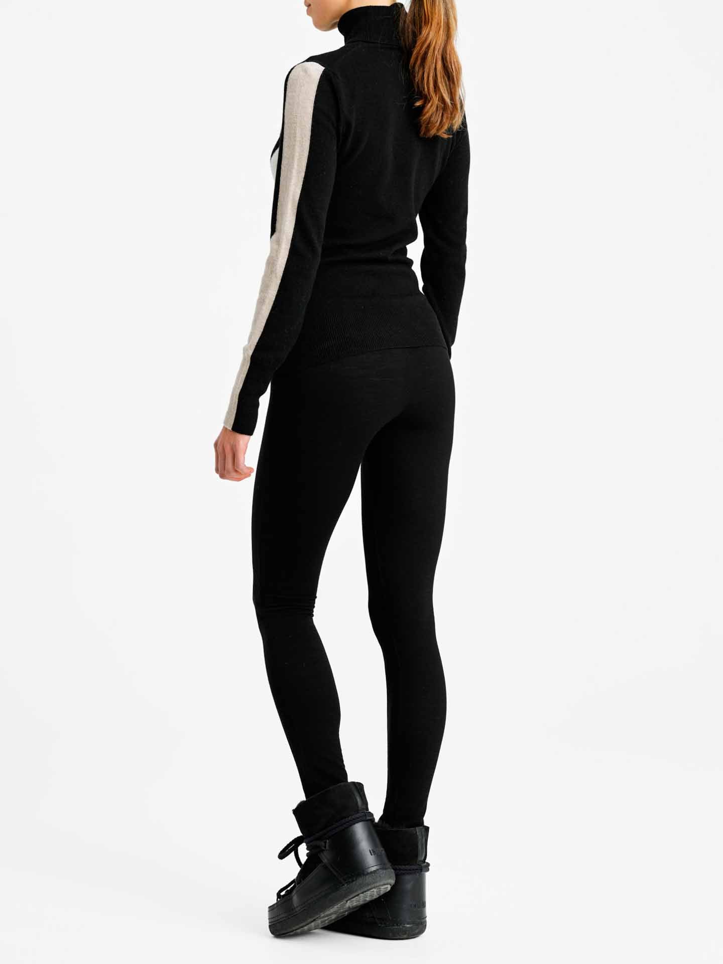 A person is standing, facing away from the camera, wearing a fitted black outfit with white accents on the sleeves. The outfit includes a long-sleeved top and We Norwegians' Ski Basic Leggings Women made of merino wool, paired with black ankle boots. The background is plain white.