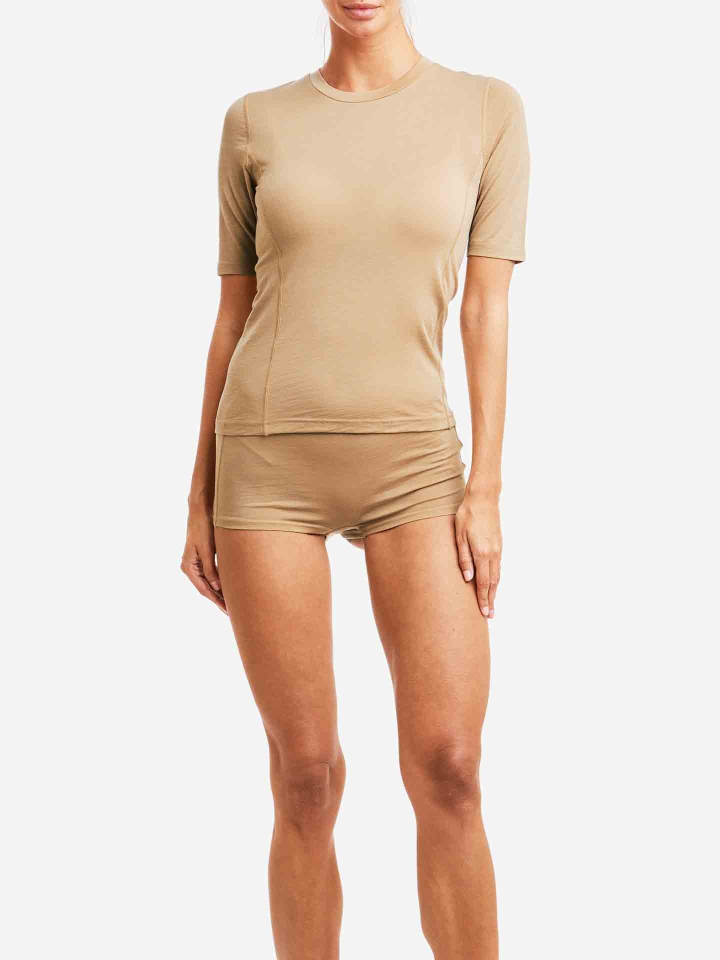 A woman is wearing the We Norwegians Sno Merino Hipster Women set, composed of a beige short-sleeve top and matching shorts made from breathable fabric. The form-fitting Scandinavian Luxury base-layers highlight her silhouette as she stands against a plain white background. Her face is not visible in the image.