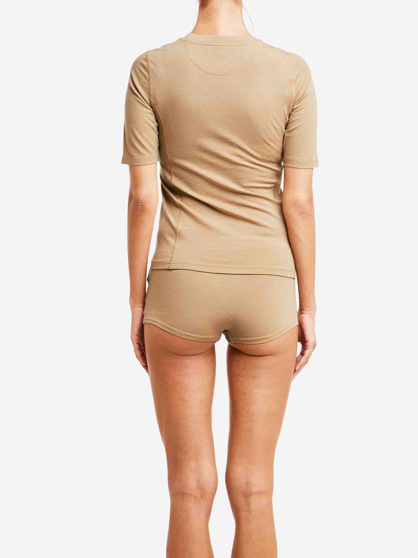 A person stands facing away from the camera, donning the We Norwegians Sno Merino Hipster Women set consisting of a beige fitted t-shirt and matching shorts made from breathable fabric. The simplicity of the plain white background accentuates the Scandinavian luxury base-layers they're wearing.