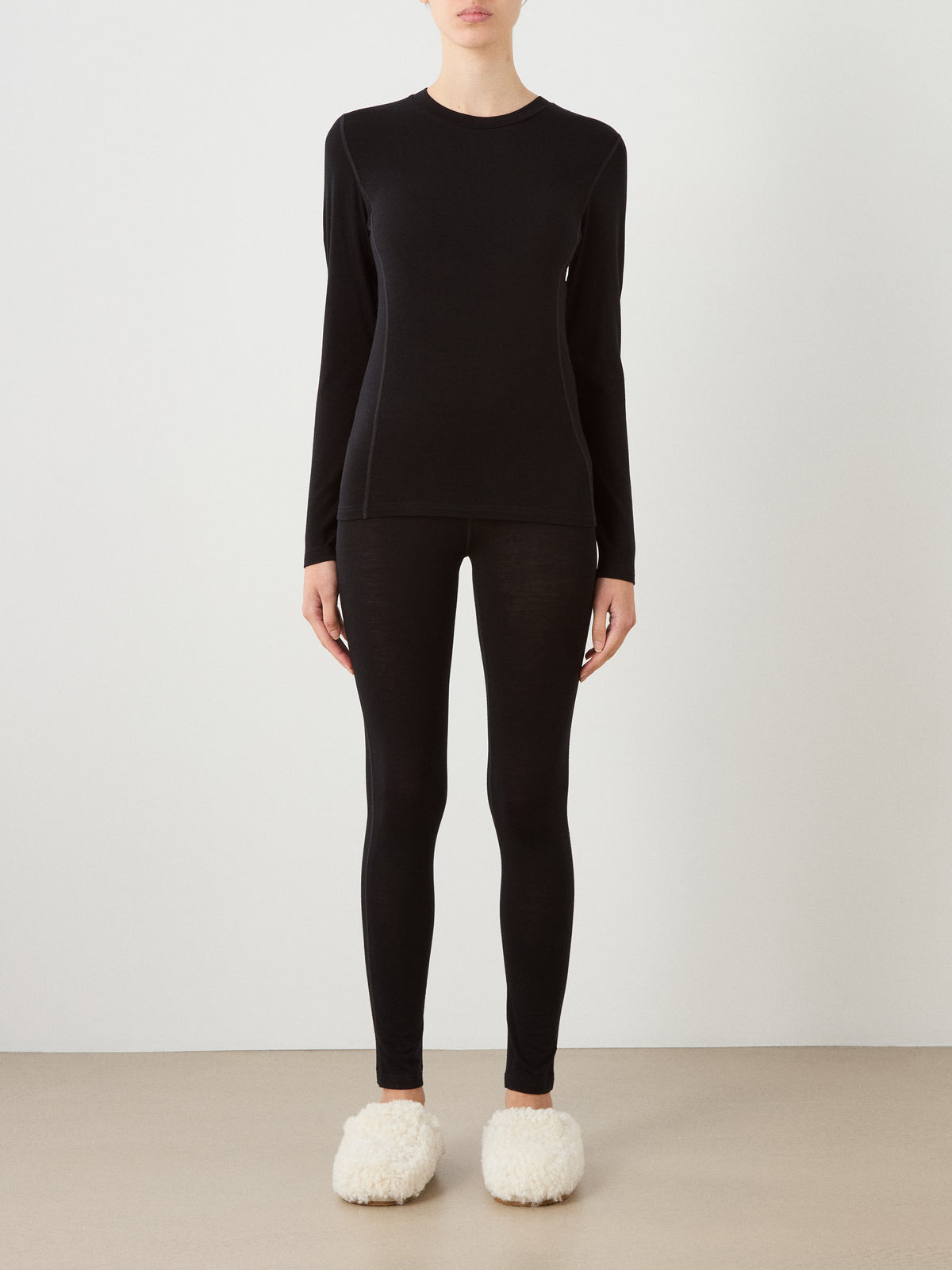 A person stands against a white wall wearing the We Norwegians Sno Merino Crewneck Women in black, paired with black leggings and fuzzy white slippers. The face is not visible in the image.