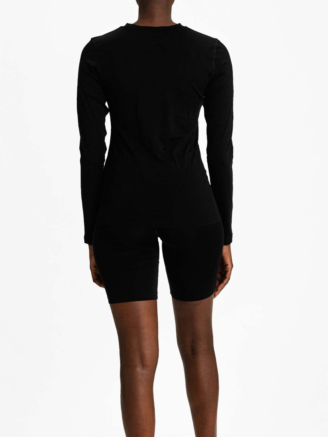 A person with dark skin is seen from the back, wearing a long-sleeved black Sno Merino Crewneck Women shirt and black fitted shorts made of super fine merino wool by We Norwegians. The plain white background highlights the Scandinavian luxury base-layers' minimalist elegance.