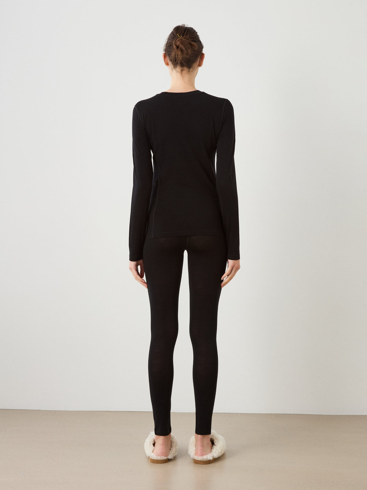 A person faces away from the camera in a black We Norwegians Sno Merino Crewneck Women and leggings, plus white fuzzy slippers, against a plain light-colored background.