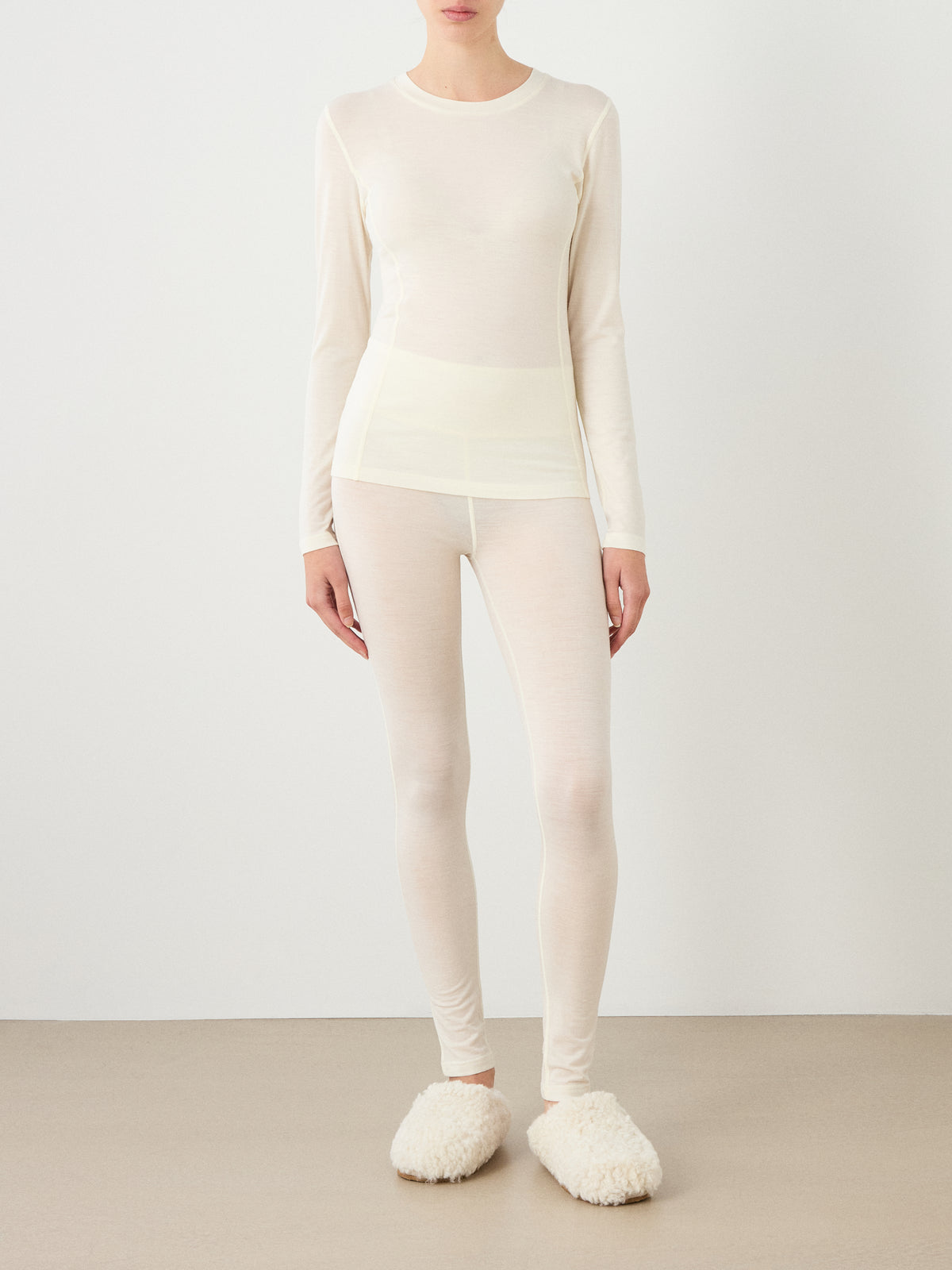 A person stands against a plain wall, wearing the We Norwegians Sno Merino Crewneck Women—a sheer, cream long-sleeve top with matching breathable leggings and fluffy cream slippers. Their face is not visible.