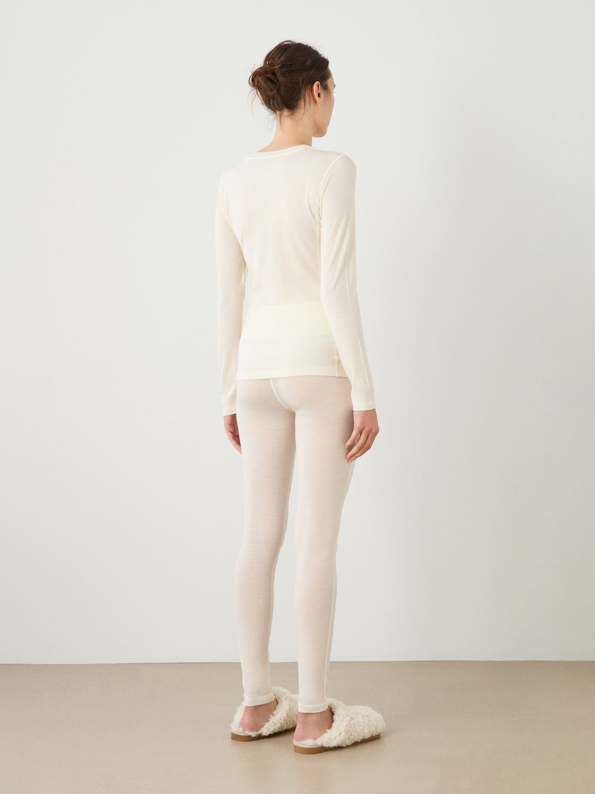 A woman stands facing away, wearing the We Norwegians Sno Merino Crewneck Women top and matching leggings in light beige with fluffy slippers, against a plain wall and neutral floor.