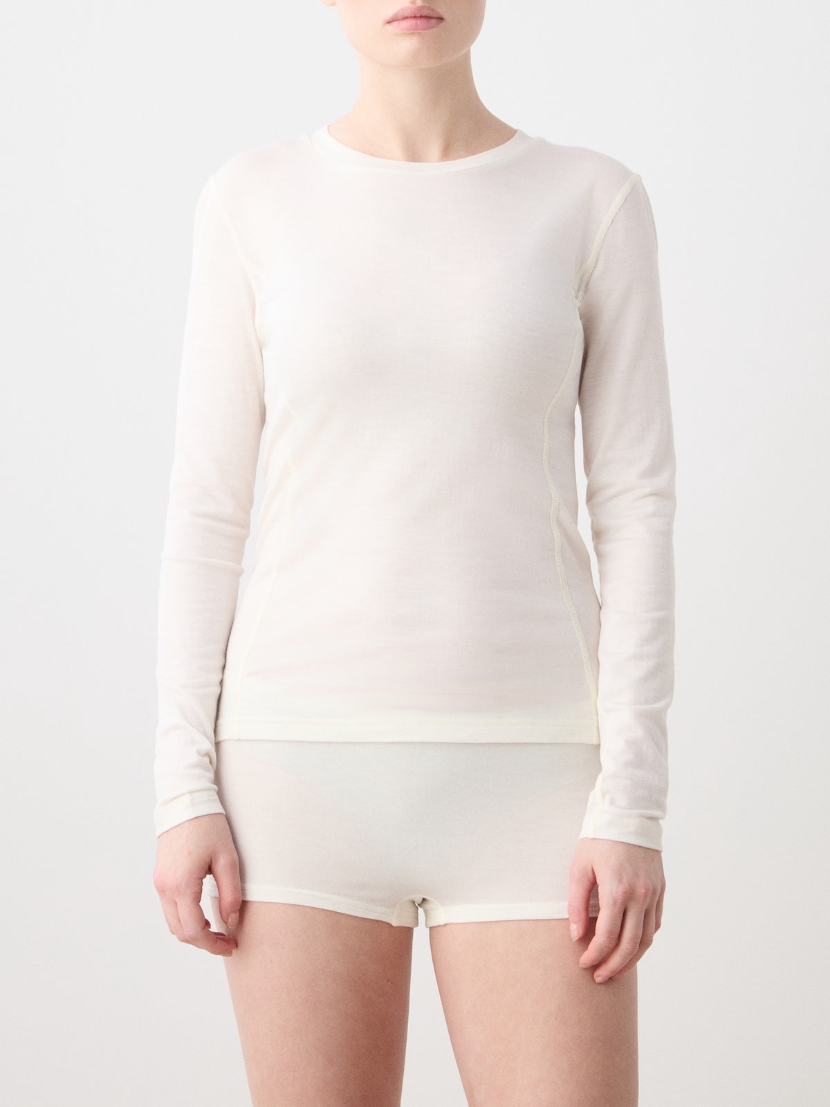 A person wearing the We Norwegians Sno Merino Crewneck Women—a long-sleeve, off-white top made from breathable single jersey—paired with matching shorts, stands against a plain, light background. The person’s face is partially out of frame.