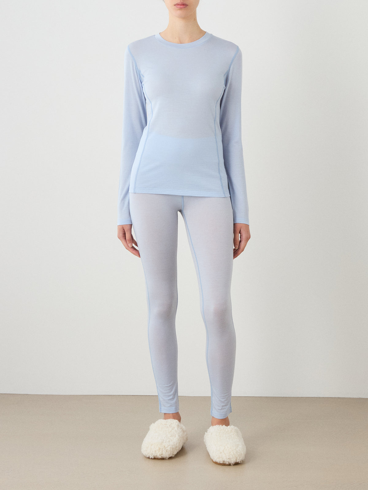 A person stands against a plain background wearing a light blue We Norwegians Sno Merino Crewneck Women and white fuzzy slippers. Their face is not visible.