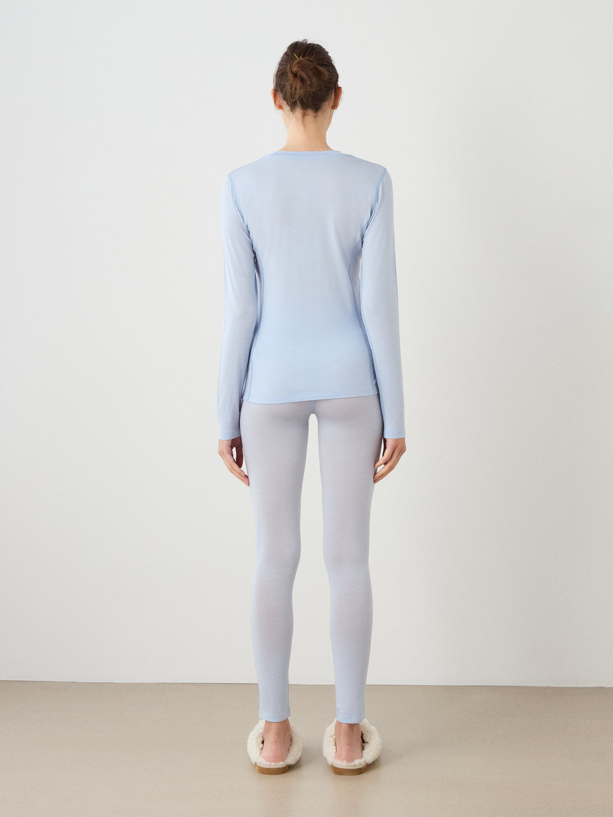 A person stands facing away in a light blue We Norwegians Sno Merino Crewneck Women top and matching leggings, both made from breathable jersey fabric, with fluffy slippers, against a white wall and beige floor.