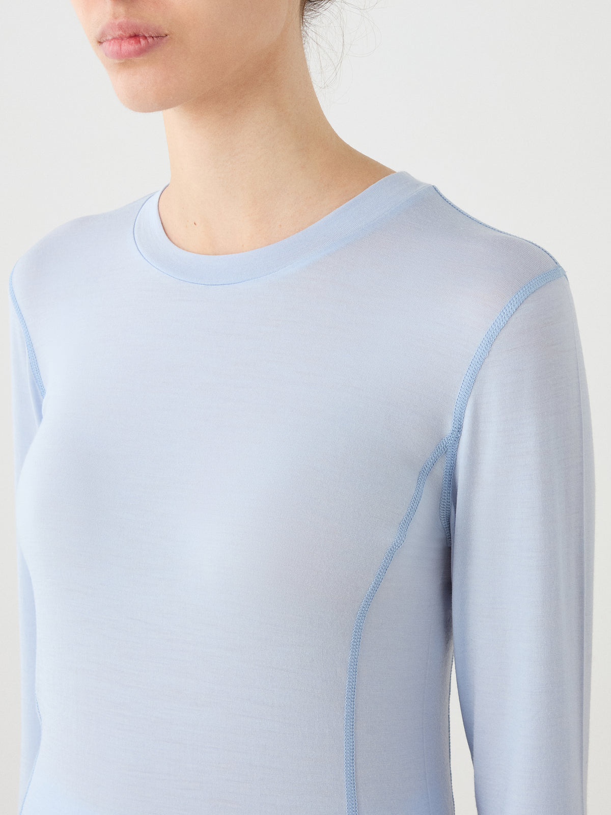 A model wears the We Norwegians Sno Merino Crewneck Women in light blue, shown from shoulders to upper chest, facing slightly left against a plain, light background.