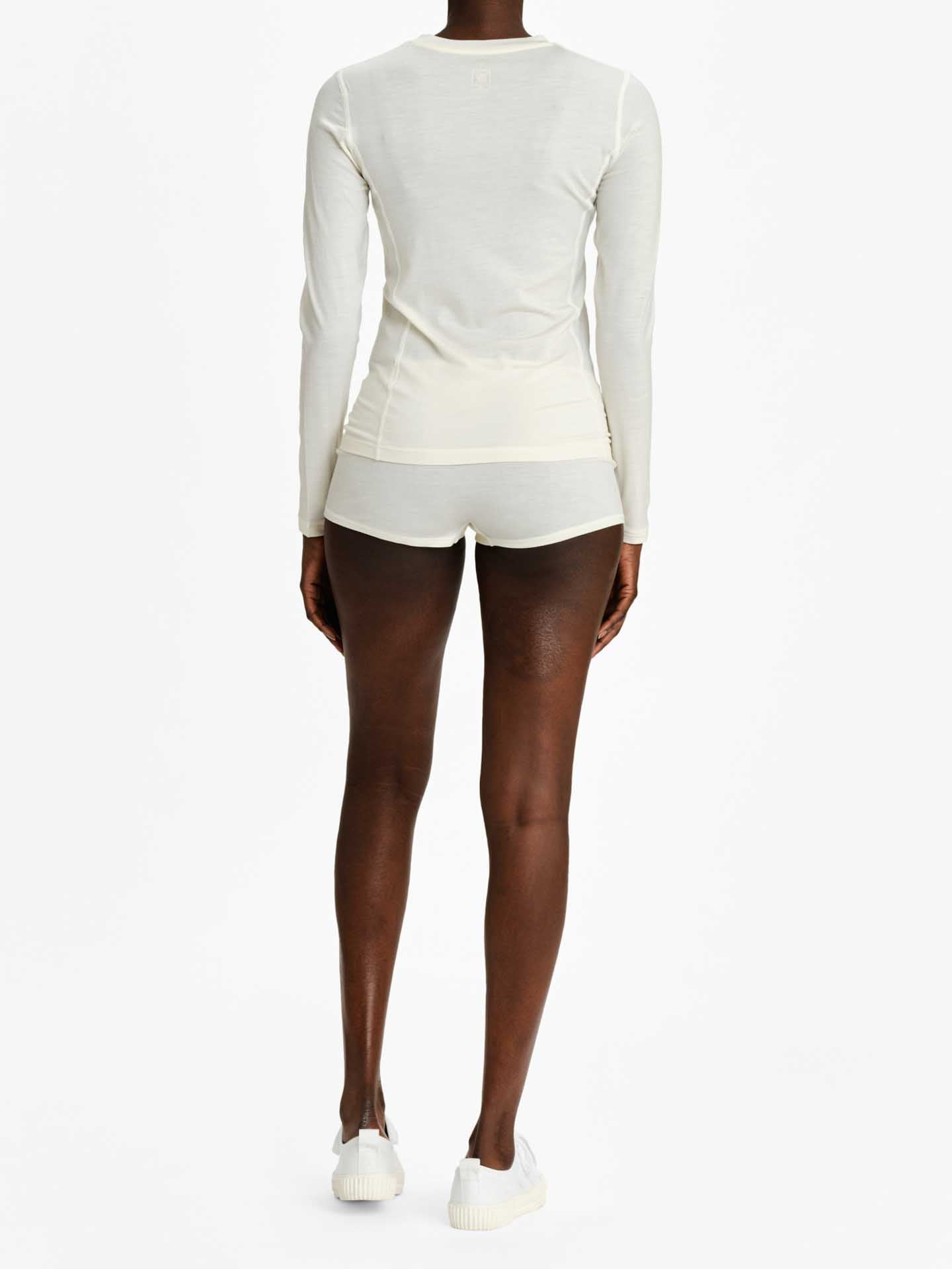 A person stands facing away from the camera, wearing a We Norwegians Sno Merino Crewneck Women in ivory, with matching shorts and white sneakers. The plain white backdrop accentuates the Scandinavian luxury base-layers of the outfit.