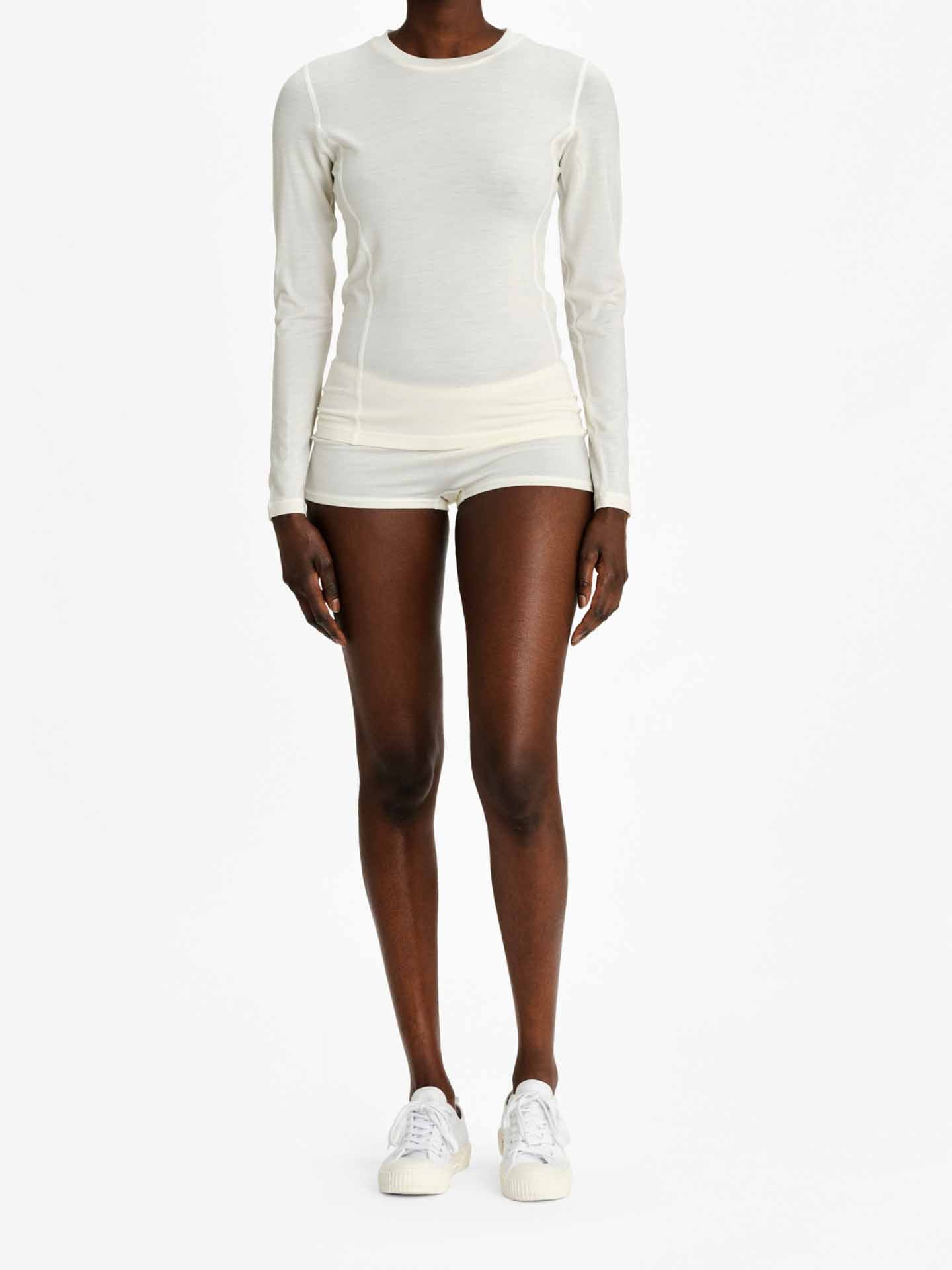 A person is standing against a plain background wearing the Sno Merino Crewneck Women by We Norwegians, along with matching white shorts and sneakers. The outfit, crafted from Creora-Eco-Soft fabric, appears to be form-fitting and designed for active wear.