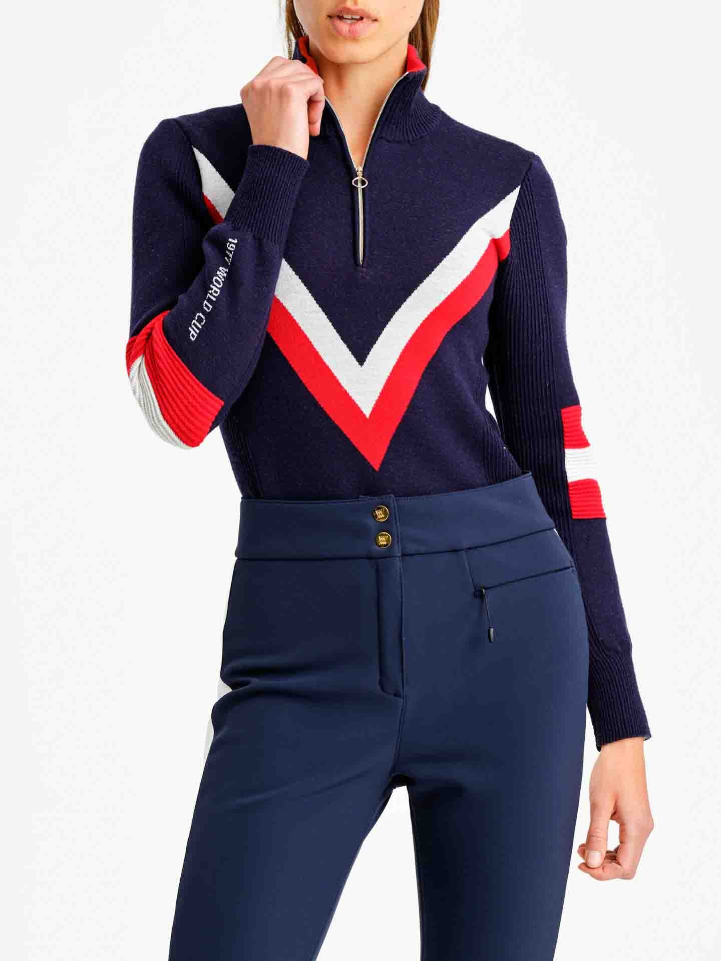 A person wearing the Hafjell Zip Up Women pullover ski sweater by We Norwegians, characterized by its navy blue and red V-shaped pattern and zip-up collar. The left sleeve boasts white and red bands, while the right features text. Meticulously crafted from fine merino wool by Italian knitters, they pair it with high-waisted navy blue pants featuring double buttons.