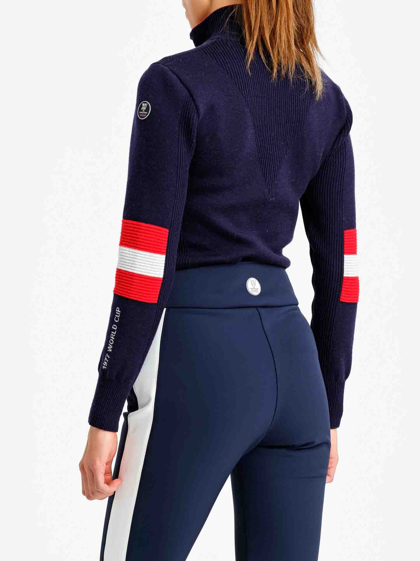 A person with long hair stands facing away, wearing the Hafjell Zip Up Women from We Norwegians— a navy blue sweater adorned with red and white bands on the sleeves, paired with navy blue pants featuring a white stripe down the sides. This fine merino wool sweater, meticulously crafted by Italian knitters, showcases a high collar and a logo patch on the upper arm.