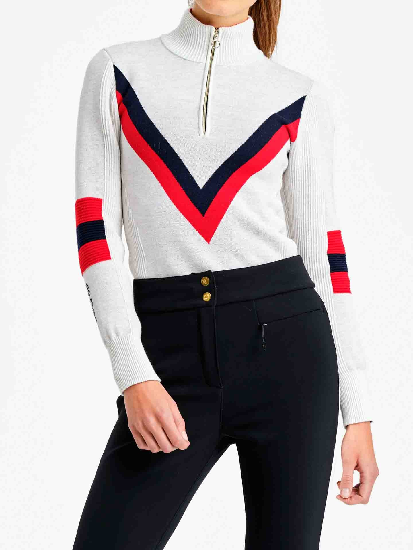 A person is wearing the Hafjell Zip Up Women by We Norwegians. This white long-sleeve pullover features a turtleneck and a half-zip closure. Crafted from fine merino wool by Italian knitters, the ski sweater showcases bold red and black chevrons on the chest along with red and black bands on the sleeves. The individual is also wearing fitted black pants.