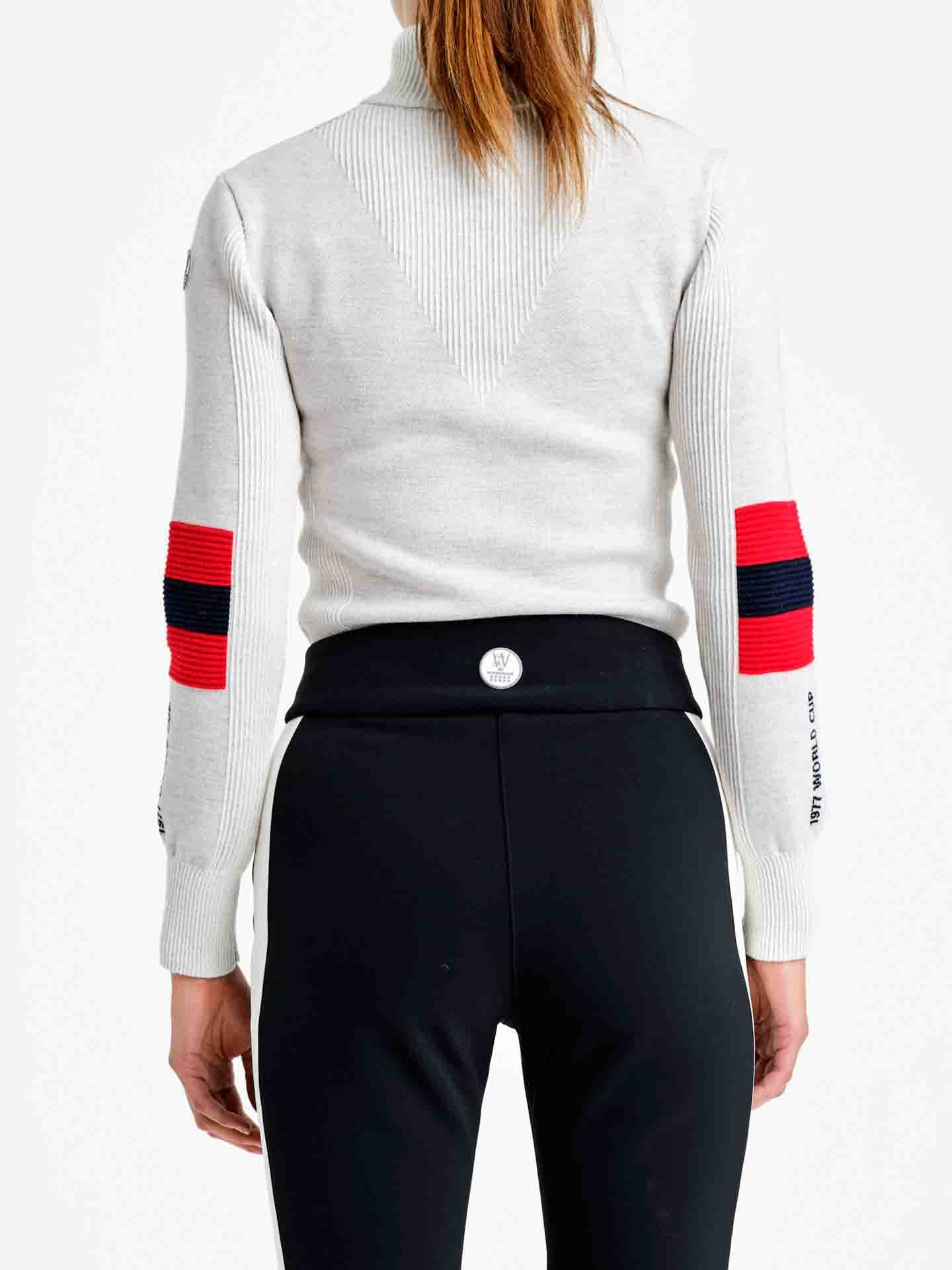 Rear view of a person wearing the We Norwegians Hafjell Zip Up Women, a white, long-sleeve ski sweater featuring red and navy blue elbow stripes, paired with black fitted pants. The person’s hair is styled in a ponytail against a plain white background. This elegant piece highlights the timeless craftsmanship of Italian knitters, crafted from fine merino wool.