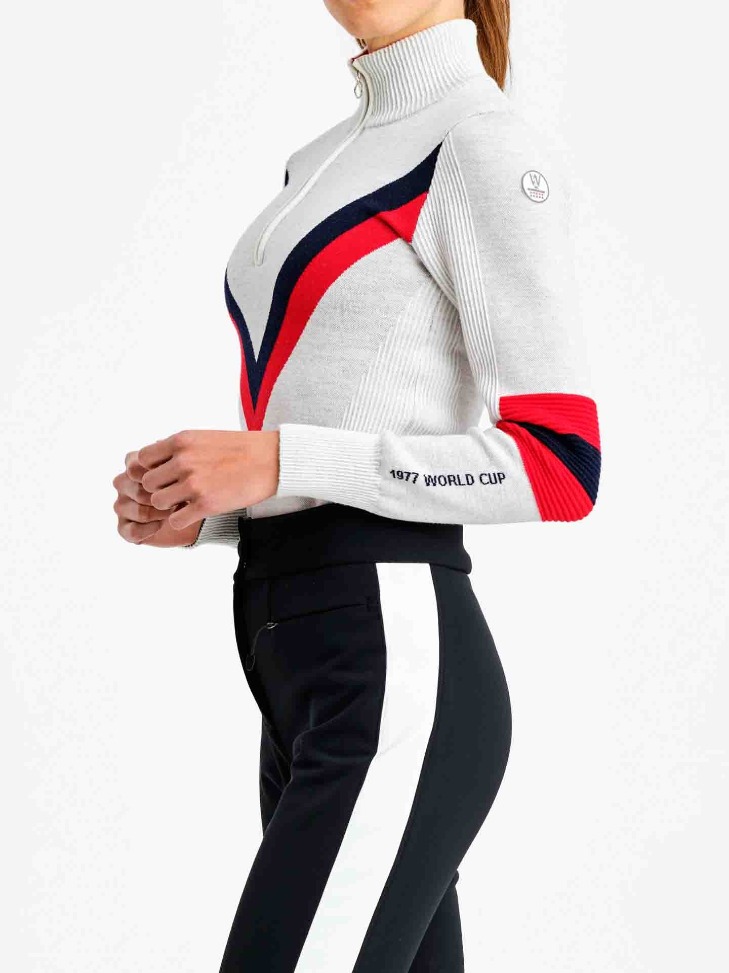 A person wearing the Hafjell Zip Up Women by We Norwegians, a stylish and fitted sweater with a high collar and zippered front, crafted from fine merino wool by Italian knitters. The sweater features white, red, and navy blue chevron stripes on the chest and arms, with "1977 WORLD CUP" written on the left sleeve. The person is also wearing black pants with a white stripe.