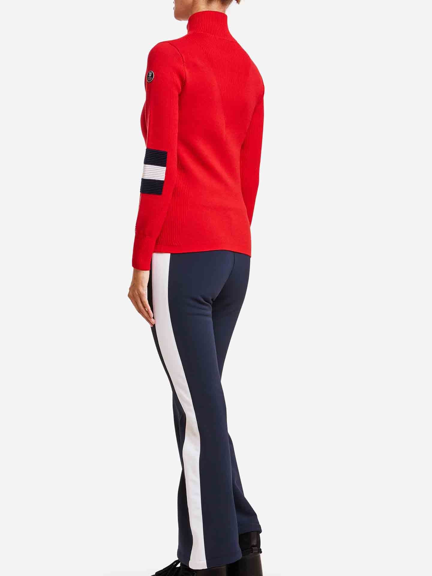 A person is standing facing away, wearing a red Hafjell Zip Up Women turtleneck sweater by We Norwegians, featuring a black and white stripe on the left sleeve, paired with navy blue pants with white side stripes. The background is plain white.