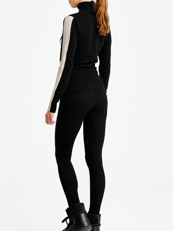 A person stands with their back to the camera, wearing the We Norwegians 1977 World Cup Sweater Women in black, featuring white stripes on the sleeves. They pair it with black leggings and ankle-high black boots. The plain white studio background enhances a sleek ski fashion vibe.