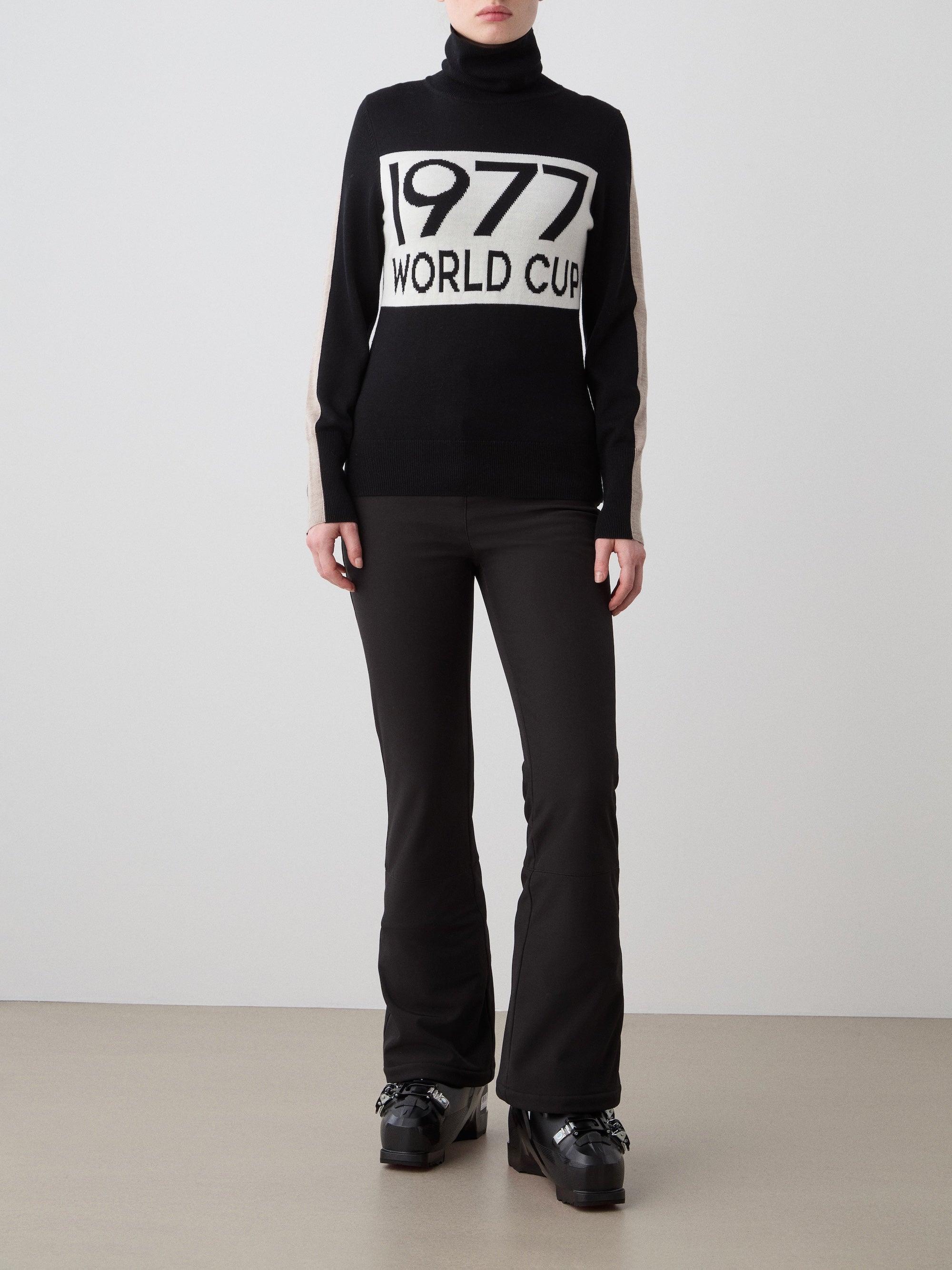 A woman wears the We Norwegians 1977 World Cup Sweater Women, inspired by 70’s ski fashion and featuring "1977 WORLD CUP" text, paired with black flared pants and platform shoes, standing against a plain background.
