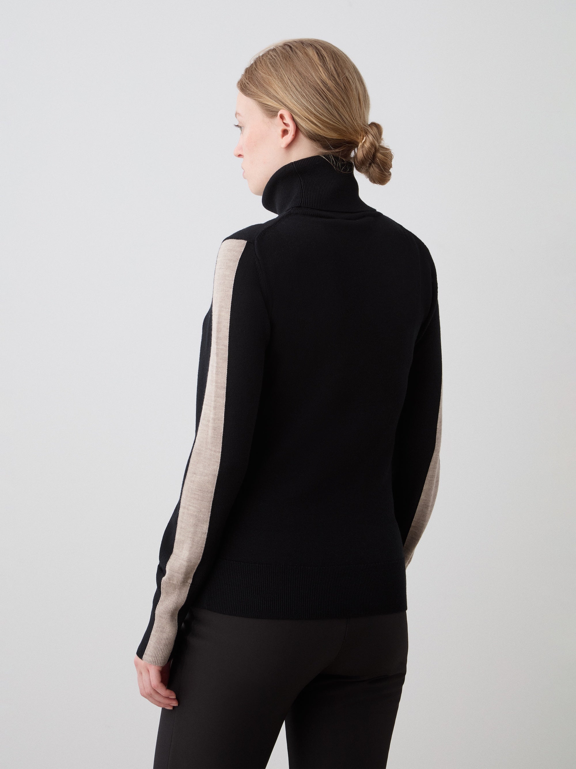 A woman with light brown hair in a low bun wears the We Norwegians 1977 World Cup Sweater Women—black with beige sleeve stripes—and black pants, channeling vintage ski style against a plain light background.