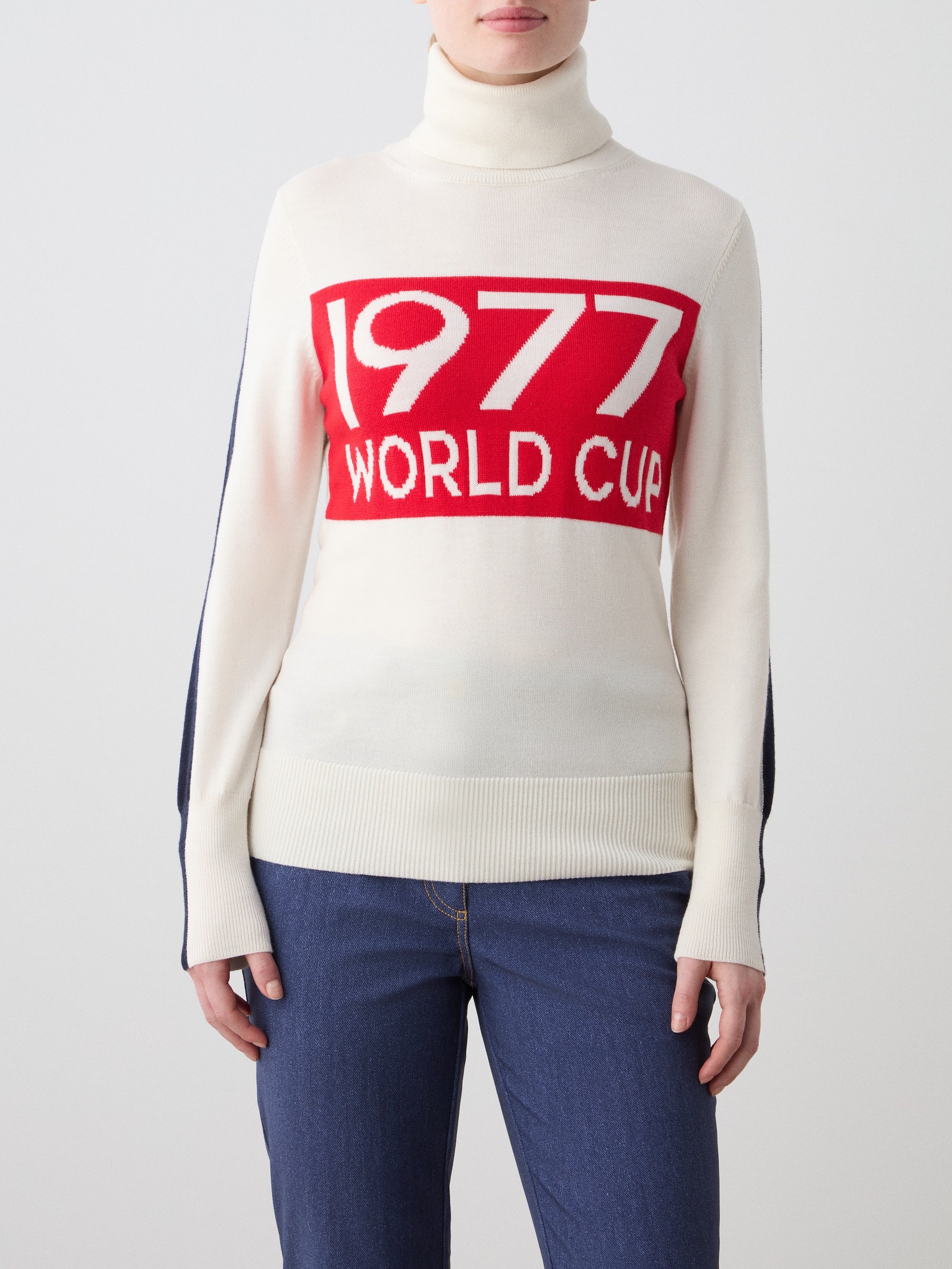 A woman wears the We Norwegians 1977 World Cup Sweater, featuring bold red "1977 WORLD CUP" lettering on cream, styled with blue jeans against a plain background—capturing classic 70’s ski fashion.