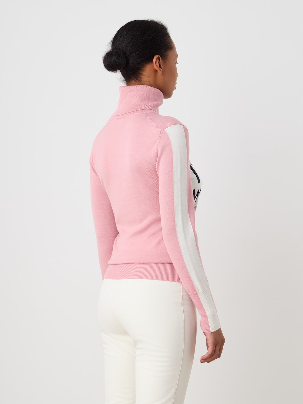 A woman with dark hair in a low bun stands against a white background, wearing We Norwegians' World Cup Sweater women - Pre Spring 26 in pink with white sleeve stripes, paired with fitted white pants.