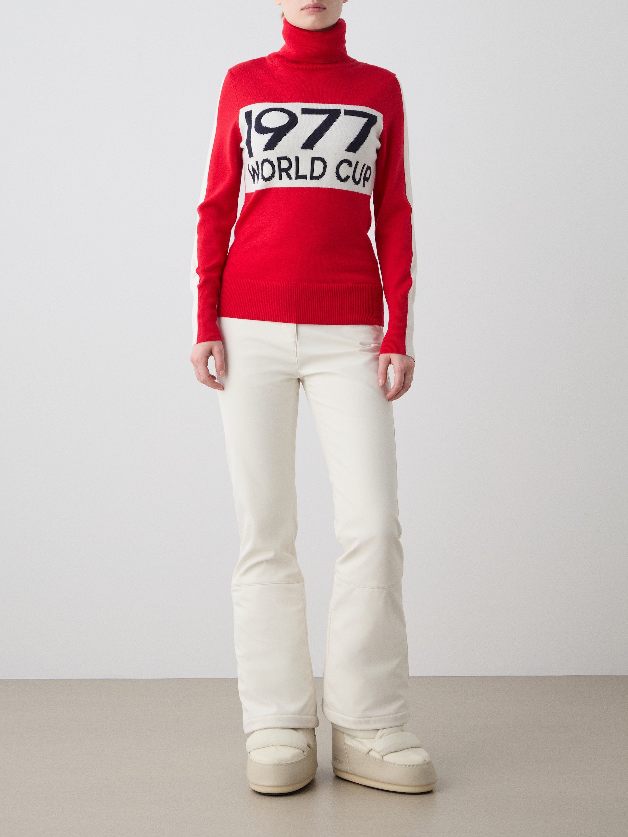 A woman faces forward in the We Norwegians 1977 World Cup Sweater—a red turtleneck with "1977 WORLD CUP" text—styled with white pants and off-white platform boots, evoking retro skiing fashion against a light backdrop.