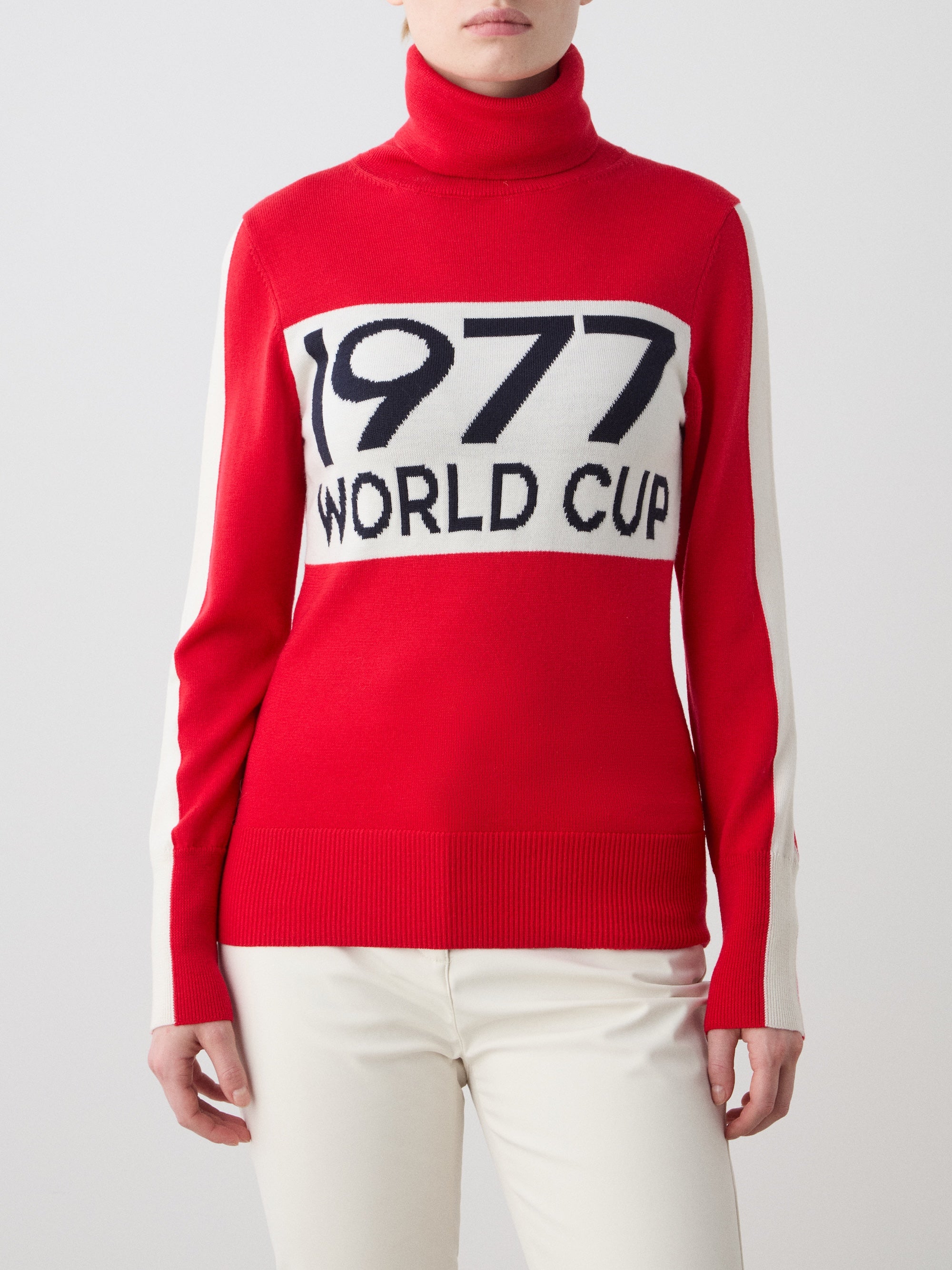 A woman wears the We Norwegians 1977 World Cup Sweater, inspired by '70s ski fashion, with a bold "1977 WORLD CUP" design and white sleeve panels, paired with white pants. Her face is partially hidden.