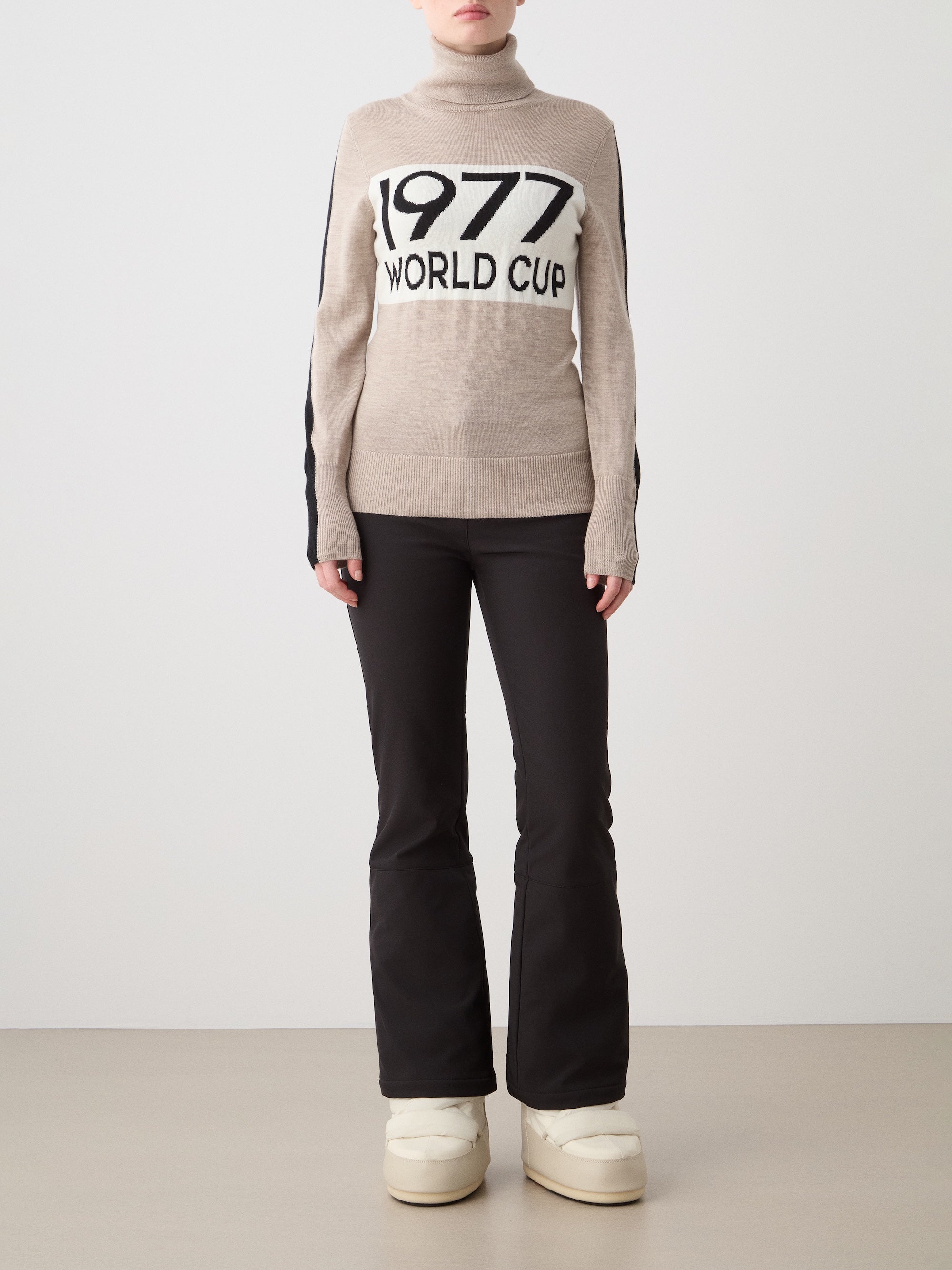 A person stands against a plain background wearing the We Norwegians 1977 World Cup Sweater Women—beige merino wool with “1977 WORLD CUP” on the front—styled with black pants and white boots; their face isn’t fully visible.