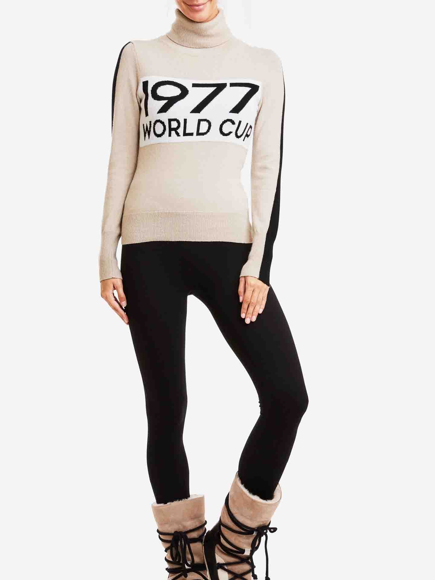 A person is dressed in the We Norwegians 1977 World Cup Sweater Women, a beige turtleneck made from merino wool with "1977 WORLD CUP" inscribed on the front. They have paired it with black leggings and are wearing brown fur-lined boots. The background is plain white.