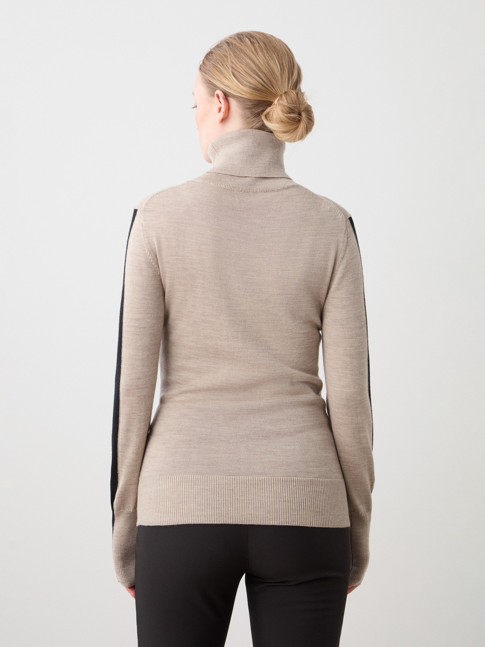 A person with blonde hair in a neat bun wears the We Norwegians 1977 World Cup Sweater Women—beige with black sleeve details—and black pants, standing back to the camera against a light background, evoking vintage ski style.