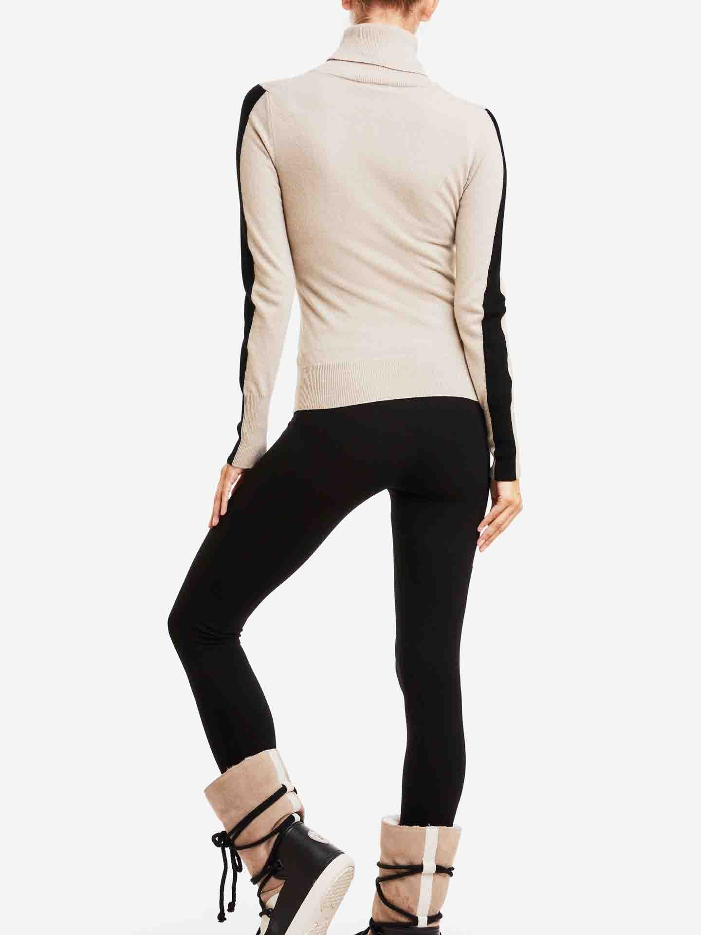 A person stands with their back turned, wearing the We Norwegians 1977 World Cup Sweater Women in beige with black sleeve accents that evoke vintage ski fashion, paired with black leggings and beige and black boots trimmed with fur. The background is plain white.