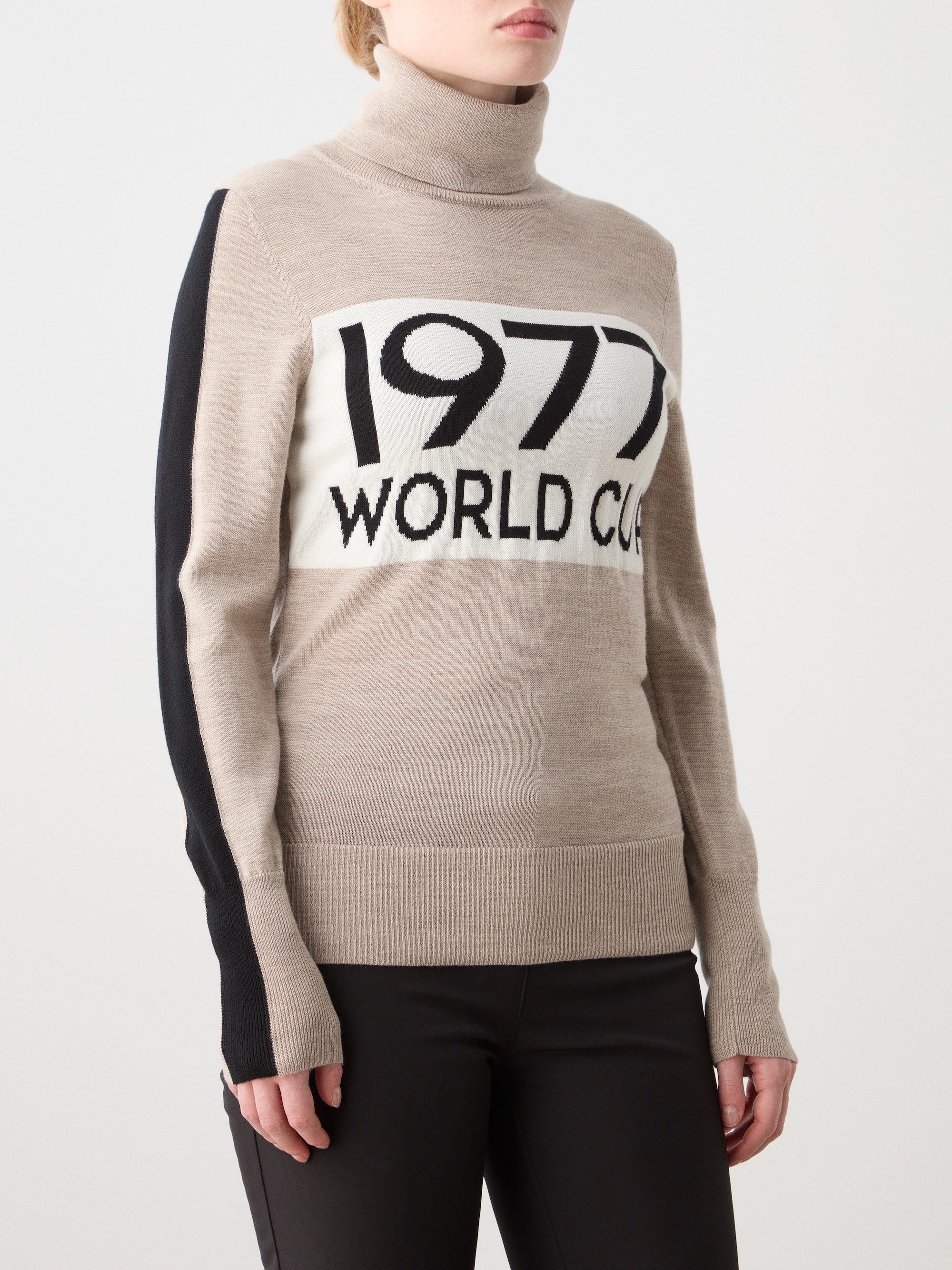 A woman wears the We Norwegians 1977 World Cup Sweater, inspired by 70s ski fashion, featuring bold "1977 WORLD CUP" text and black sleeve accents, styled with black pants.