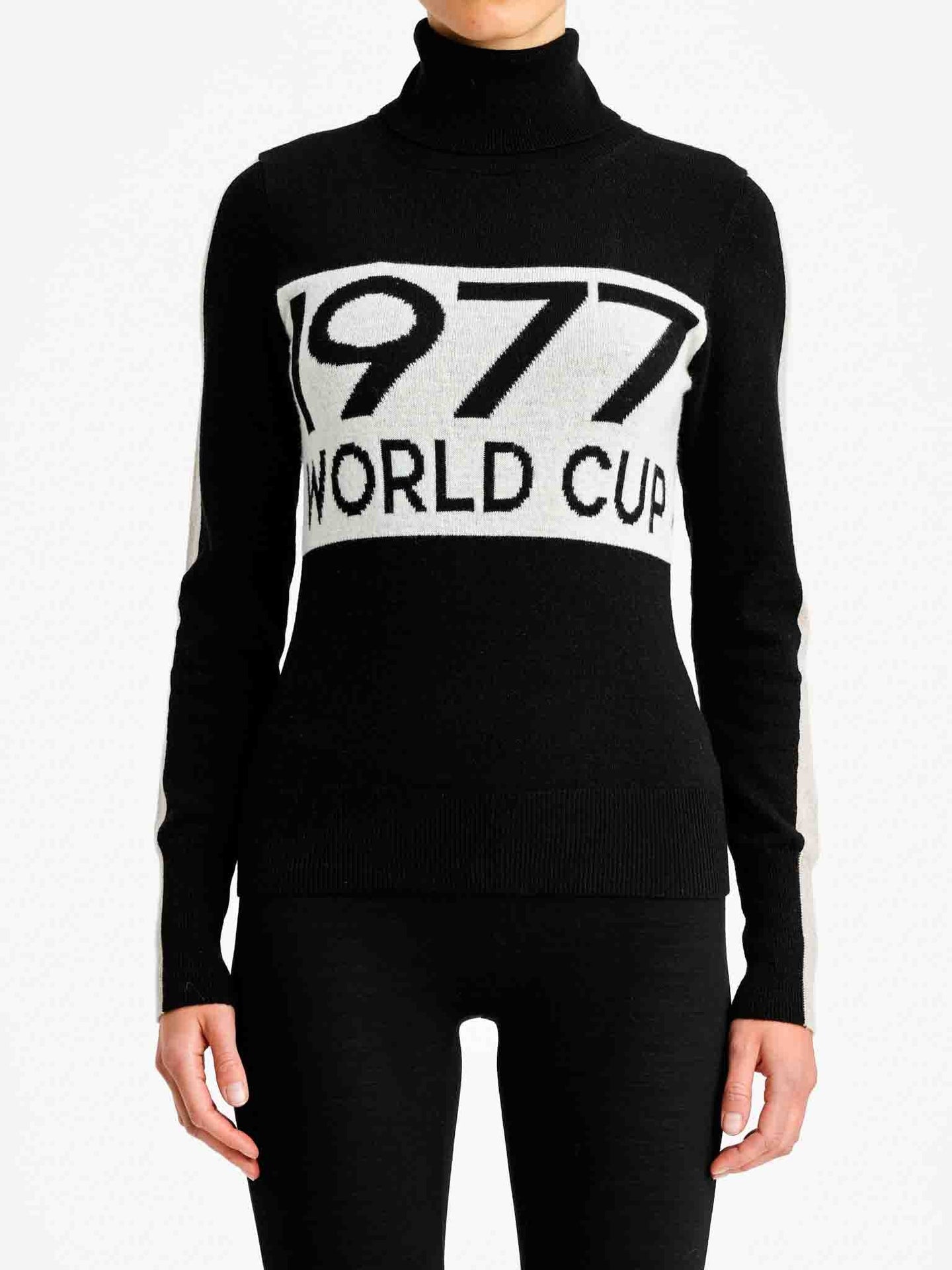 A person is wearing the "1977 World Cup Sweater Women" by We Norwegians, a stylish black and white turtleneck made from premium merino wool, with "1977 WORLD CUP" prominently displayed on the front. Paired with black pants, they stand against a plain white background, perfectly capturing the essence of vintage ski fashion.