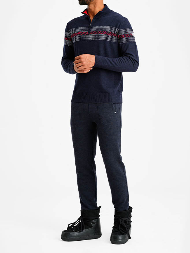 A person is standing against a plain white background, wearing the Signature Zip Up Men sweater by We Norwegians. The sweater is a long-sleeve, dark blue garment with red and white patterned design reminiscent of Norwegian heritage across the chest and shoulders. They are also wearing dark blue pants and black winter boots. The person's face is not visible.